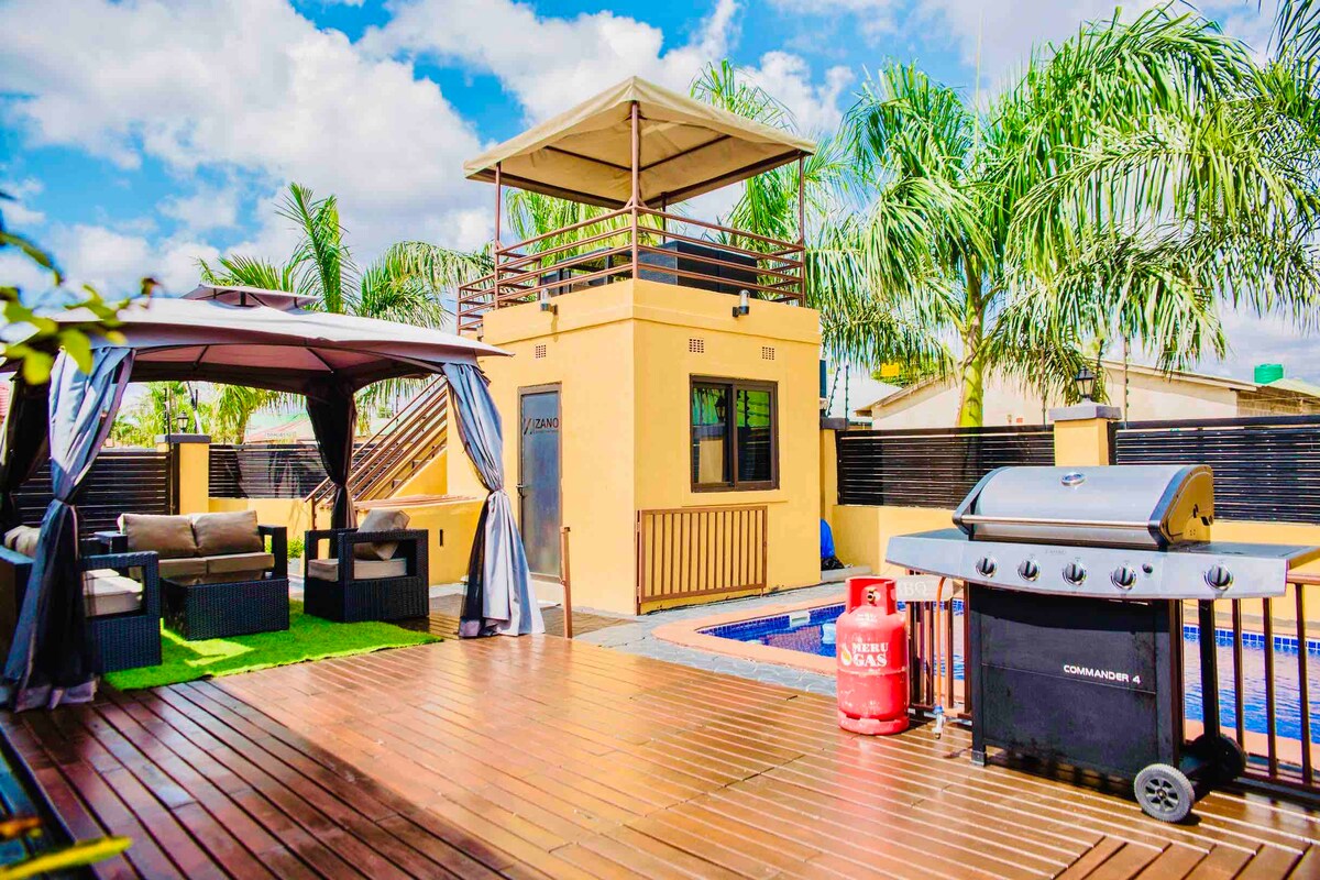 A patio area is presented with comfortable seating beneath a gazebo, surrounded by palm trees. A BBQ grill is positioned nearby, with a gas tank accessible. A roof terrace above the guard house overlooks a small pool, with wooden decking creating an inviting outdoor space.