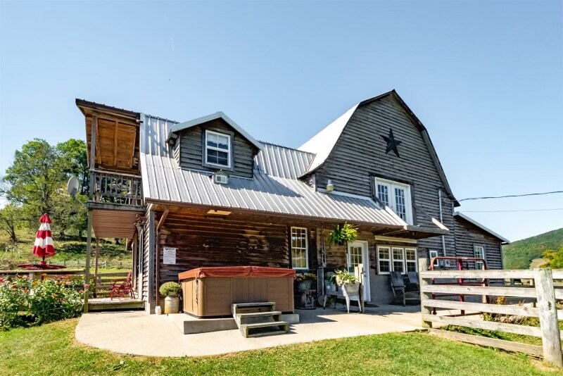 The exterior of the barn apartment showcases a rustic design with a metal roof and wooden siding. A hot tub is positioned on the patio, accompanied by potted plants and outdoor furniture. A large star decoration is prominently featured above the entrance, enhancing the farmstead charm.