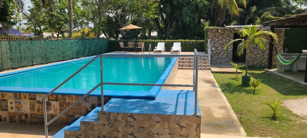 A clear blue swimming pool is framed by a stone wall, with lounge chairs positioned nearby. A shaded area features a large umbrella and additional seating. Lush greenery surrounds the space, and a hammock is visible, adding to the relaxing atmosphere of the outdoor area.