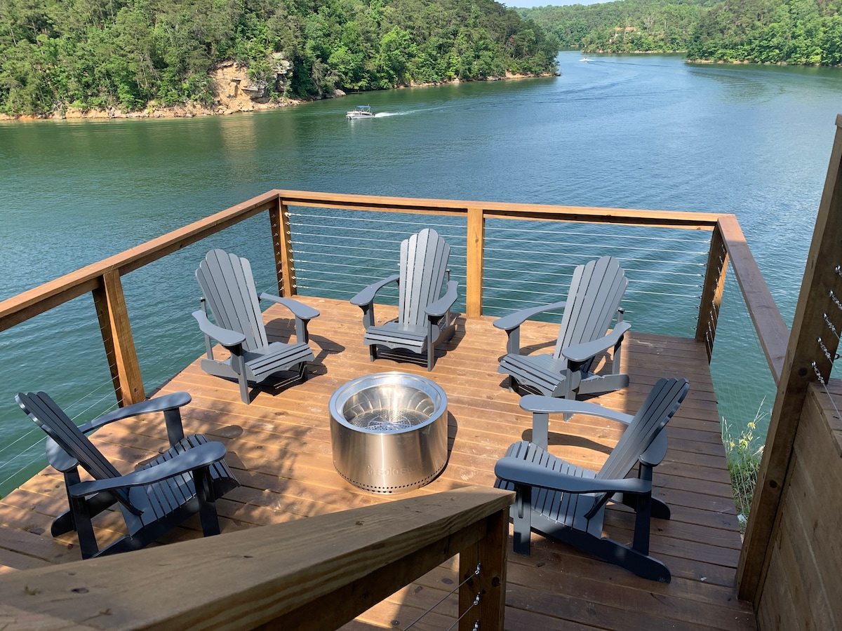 A spacious outdoor deck overlooks a serene lake, equipped with six gray adirondack chairs arranged around a central fire pit. The surrounding trees and distant shoreline are visible, enhancing the tranquil lakeside atmosphere.