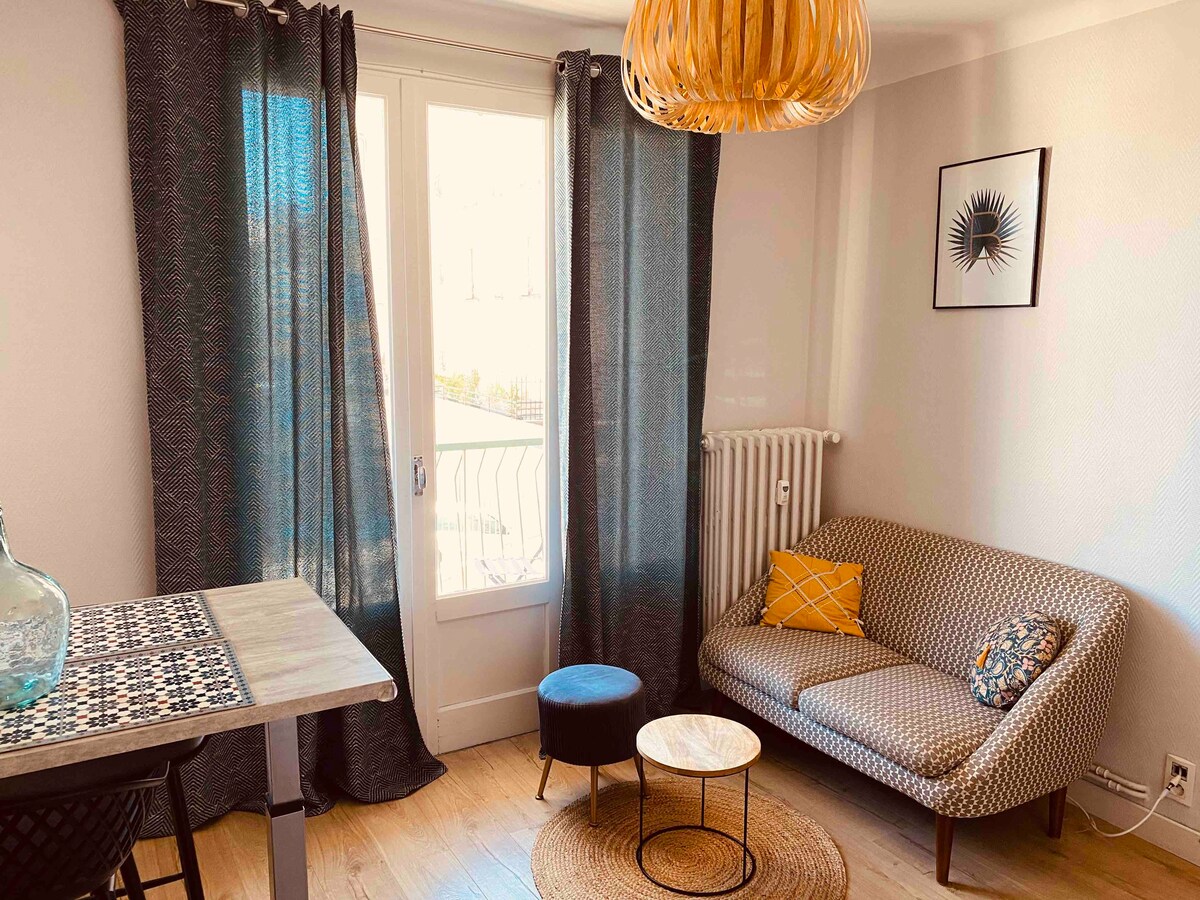 A living area features a patterned sofa with a yellow accent cushion, paired with a small black table. Natural light enters through a window dressed with dark curtains, while a round jute rug complements the wooden flooring. A minimalist wall art piece is displayed nearby.