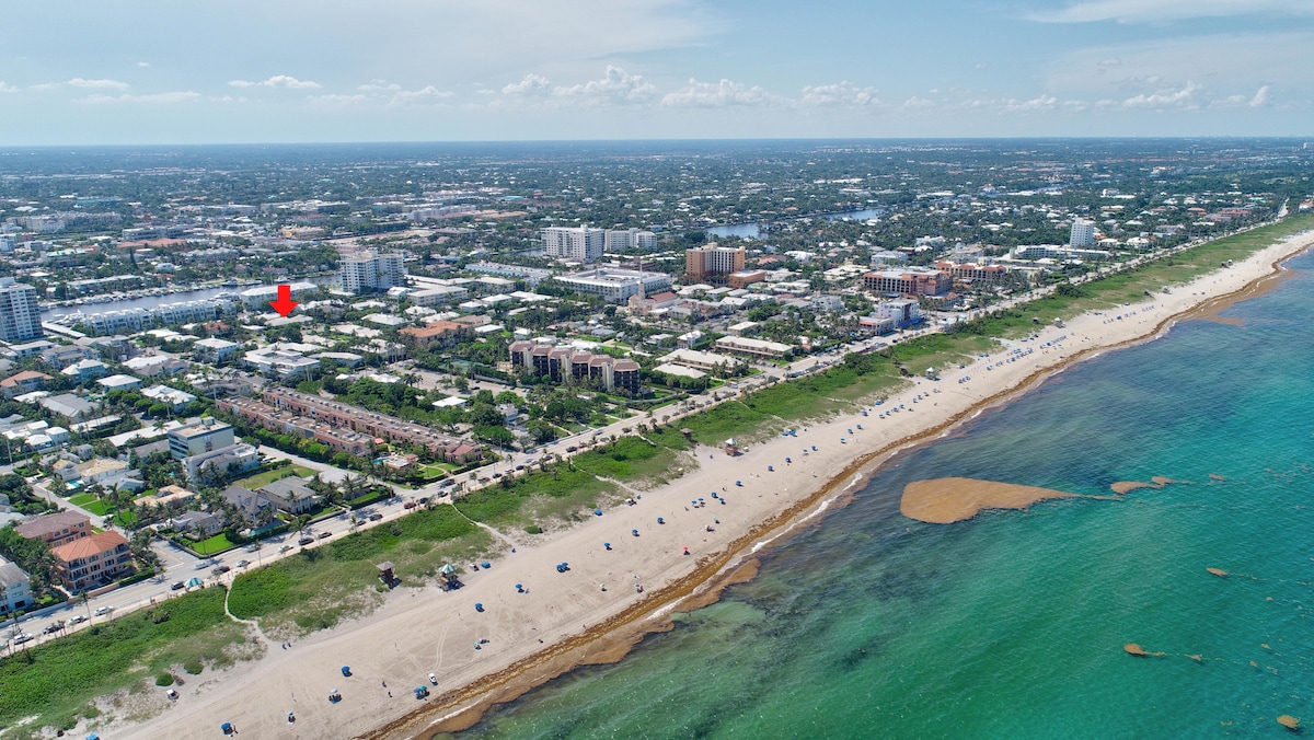 An aerial view captures the coastline and the vibrant beach area. Clear turquoise waters meet a sandy shore lined with beachgoers and umbrellas. Buildings and greenery are visible in the background, with the town's vibrant community highlighted against the expansive sky.