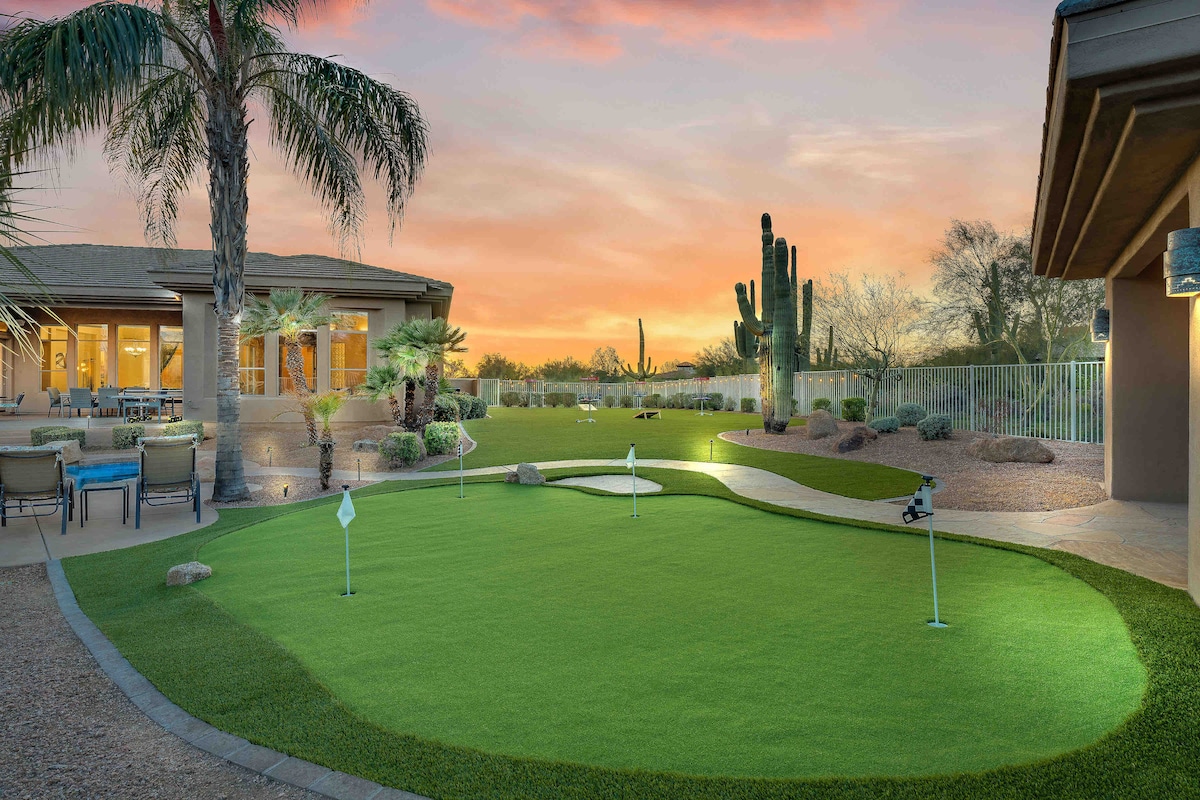 An artificial putting green features two holes against a backdrop of desert landscaping. Lush greenery contrasts with the surrounding gravel. The setting includes palm and cactus plants, with a vibrant sunset sky illuminating the scene. Cozy seating areas are visible in the distance.