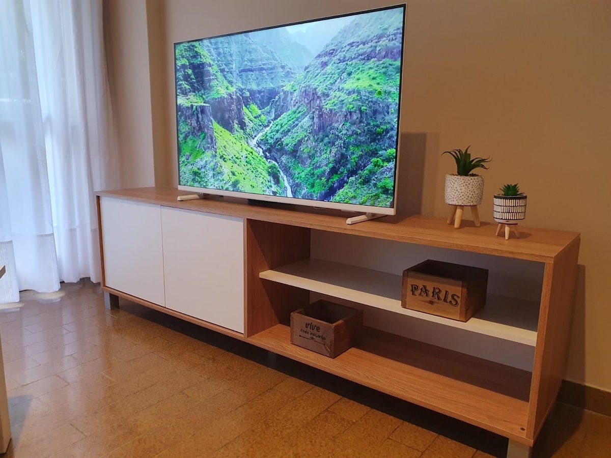 A modern entertainment unit features a large flat-screen television displaying a scenic landscape. Below the TV, open shelves host decorative plants and a few storage boxes, contributing to a clean and organized appearance in the living area.