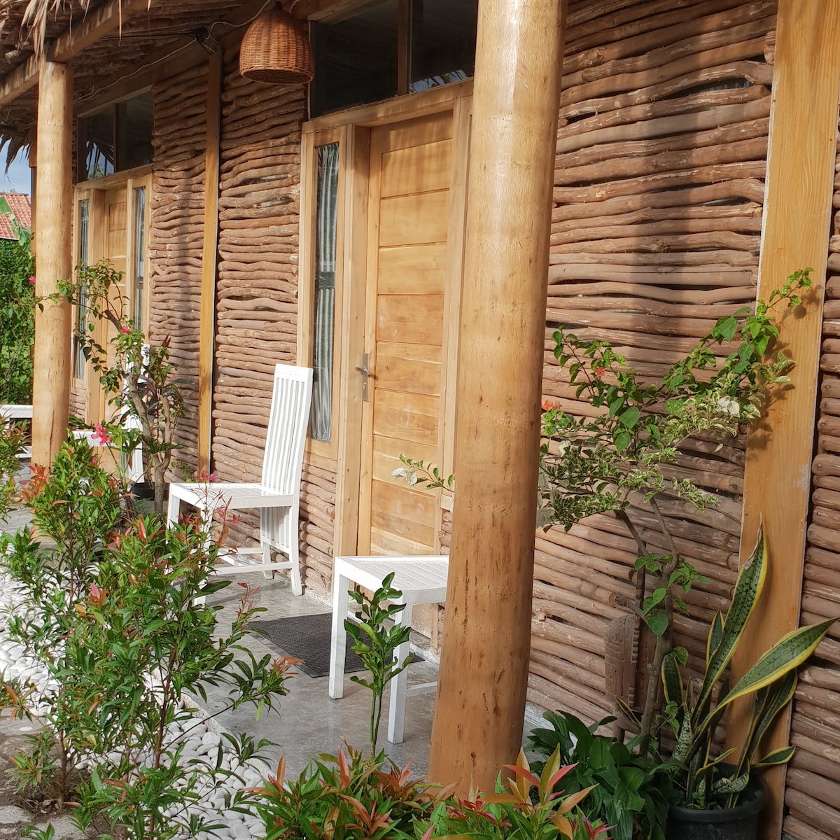 A charming exterior features a textured, natural wood facade with two wooden doors. Two white chairs are positioned beside each door, framed by potted plants and bushes that provide a touch of greenery. A stone pathway leads to the entrance.