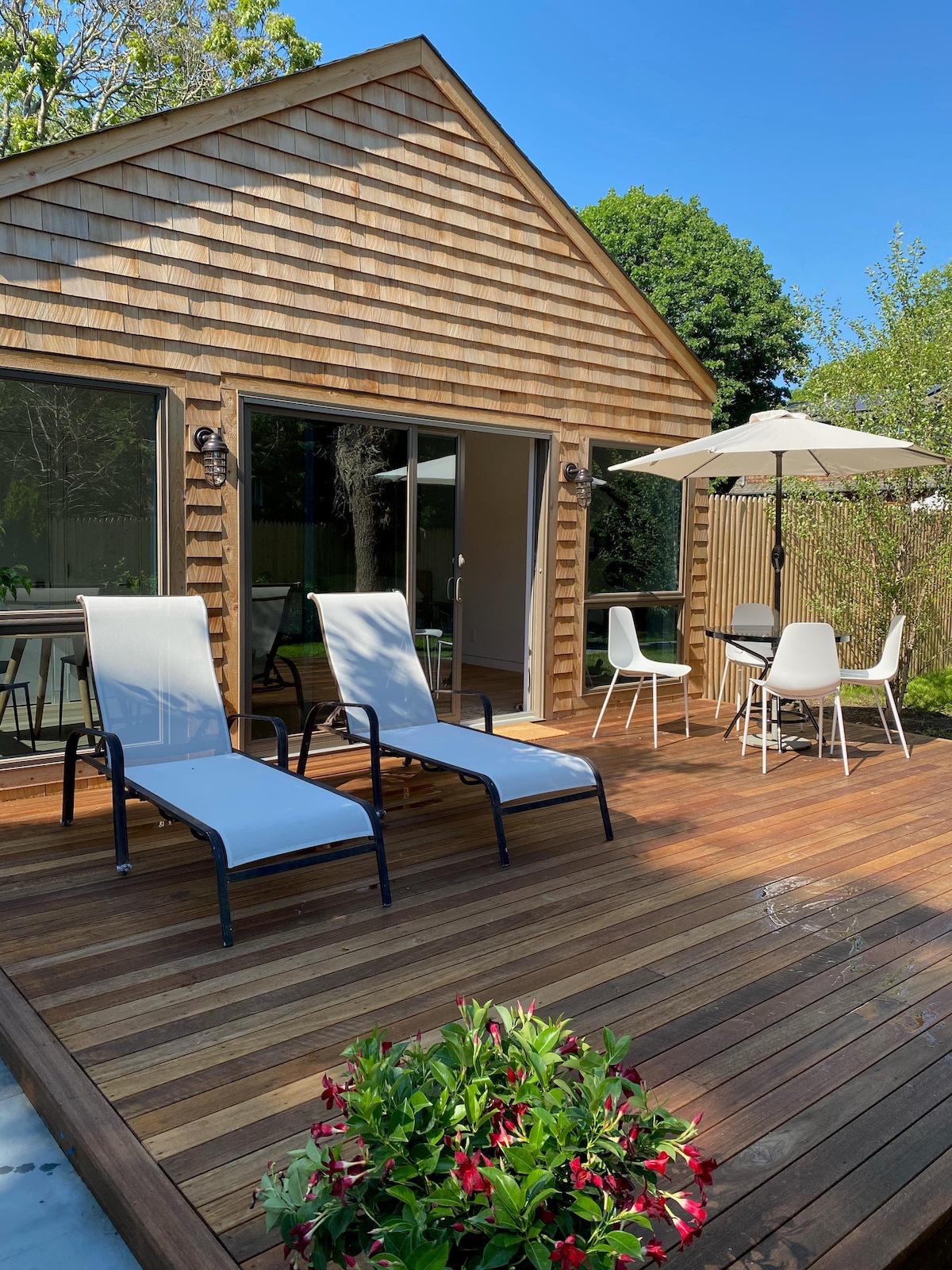 A private outdoor deck features two lounge chairs facing toward a lawn, accompanied by a round dining table with four chairs and an umbrella for shade. Surrounding greenery provides a serene setting, complemented by wooden siding on the cottage's exterior.