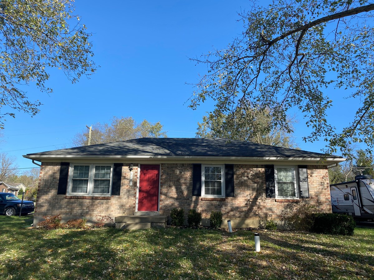 The exterior of a single-story brick home is displayed, featuring a red front door framed by two windows on each side. The light-colored siding contrasts with the lush green lawn, which is dotted with fallen leaves. A clear blue sky is visible overhead.