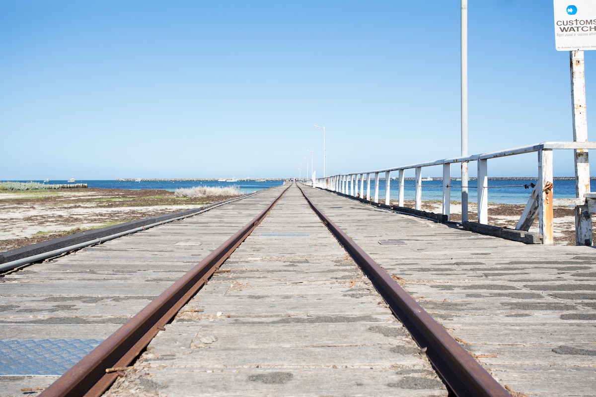 A wooden pier stretches towards the horizon, lined with rail tracks that lead to the sea. The clear blue sky complements the tranquil ocean views at both sides of the structure. Minimal distractions create a serene atmosphere, perfect for leisurely walks.