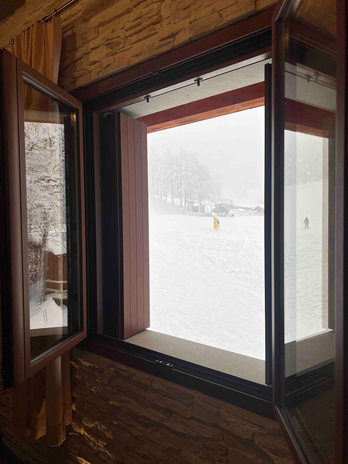 A large window reveals a snowy landscape outside, with skiers on a slope. The scene is framed by wooden window frames, and a soft haze from falling snow adds to the tranquil winter atmosphere.