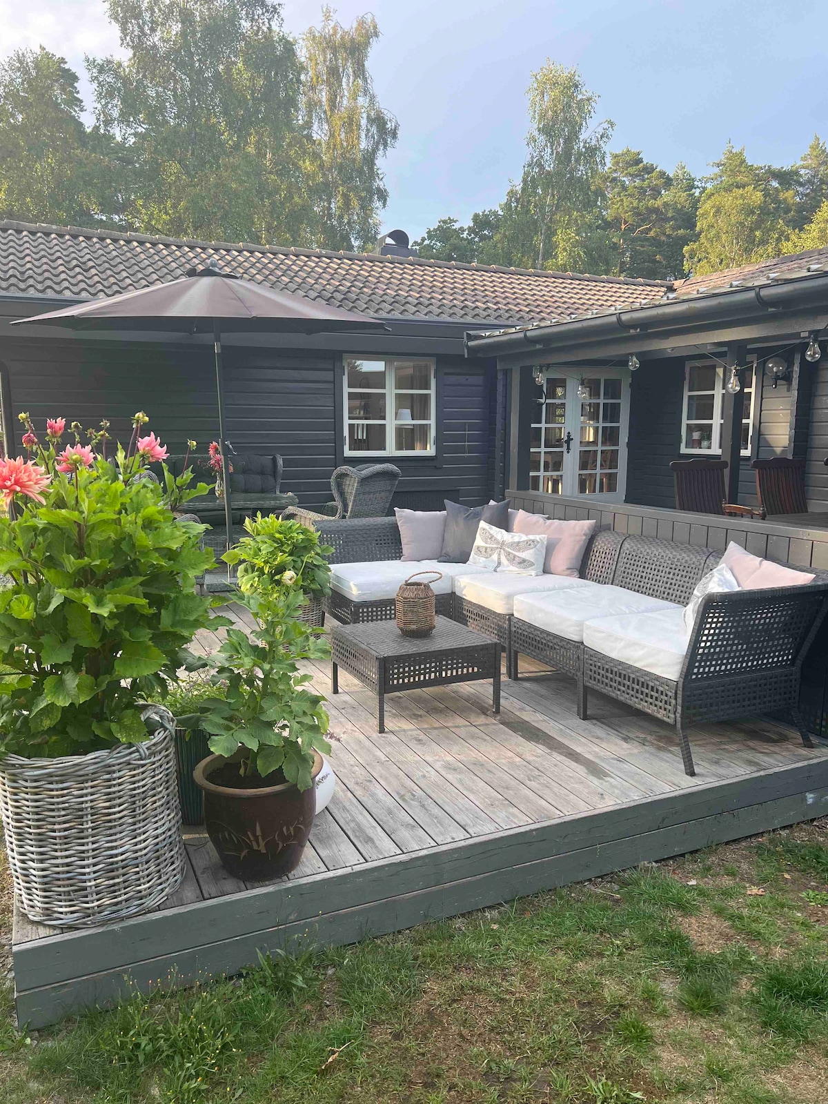 A cozy outdoor seating area is situated on a wooden deck, featuring a comfortable sectional sofa with soft cushions. Potted plants add greenery, while an umbrella provides shade, creating an inviting space for relaxation amid the natural surroundings.