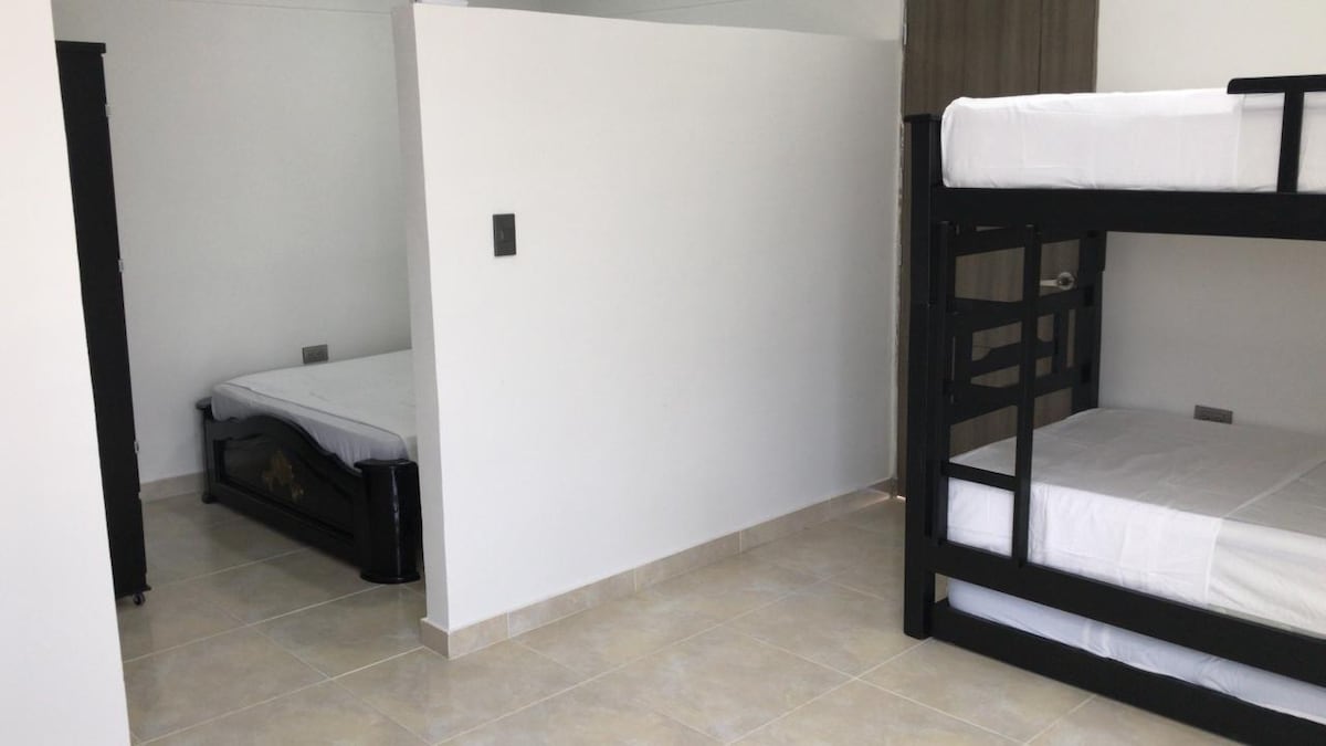 A divided sleeping area presents a double bed on one side and a two-level bunk bed on the other. The beds are neatly made with white linens. Neutral-toned walls and tiled floors create a simple and functional environment.