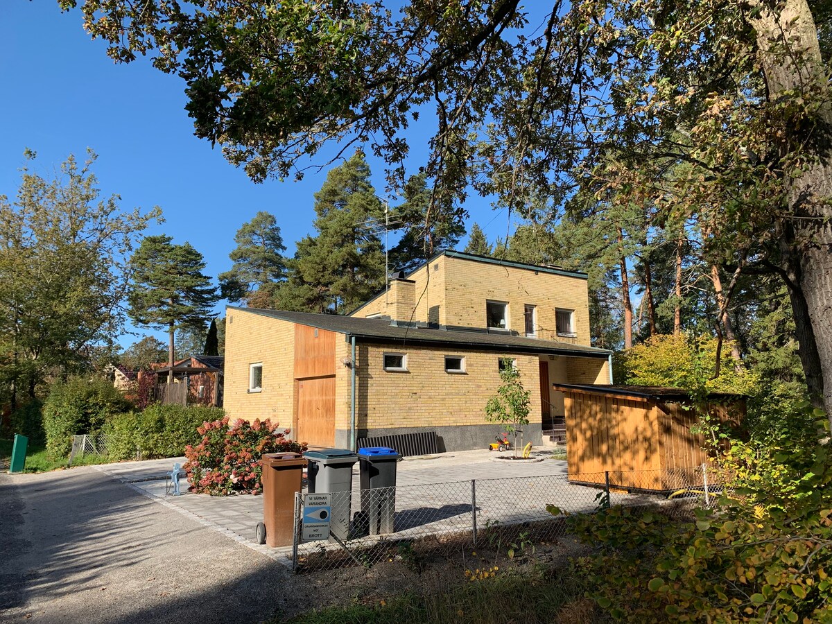 The exterior of a modern villa is displayed, featuring a warm brick facade surrounded by trees and landscaped greenery. A driveway leads to the entrance, with several bins organized at the side. The sun illuminates the area under a clear blue sky.