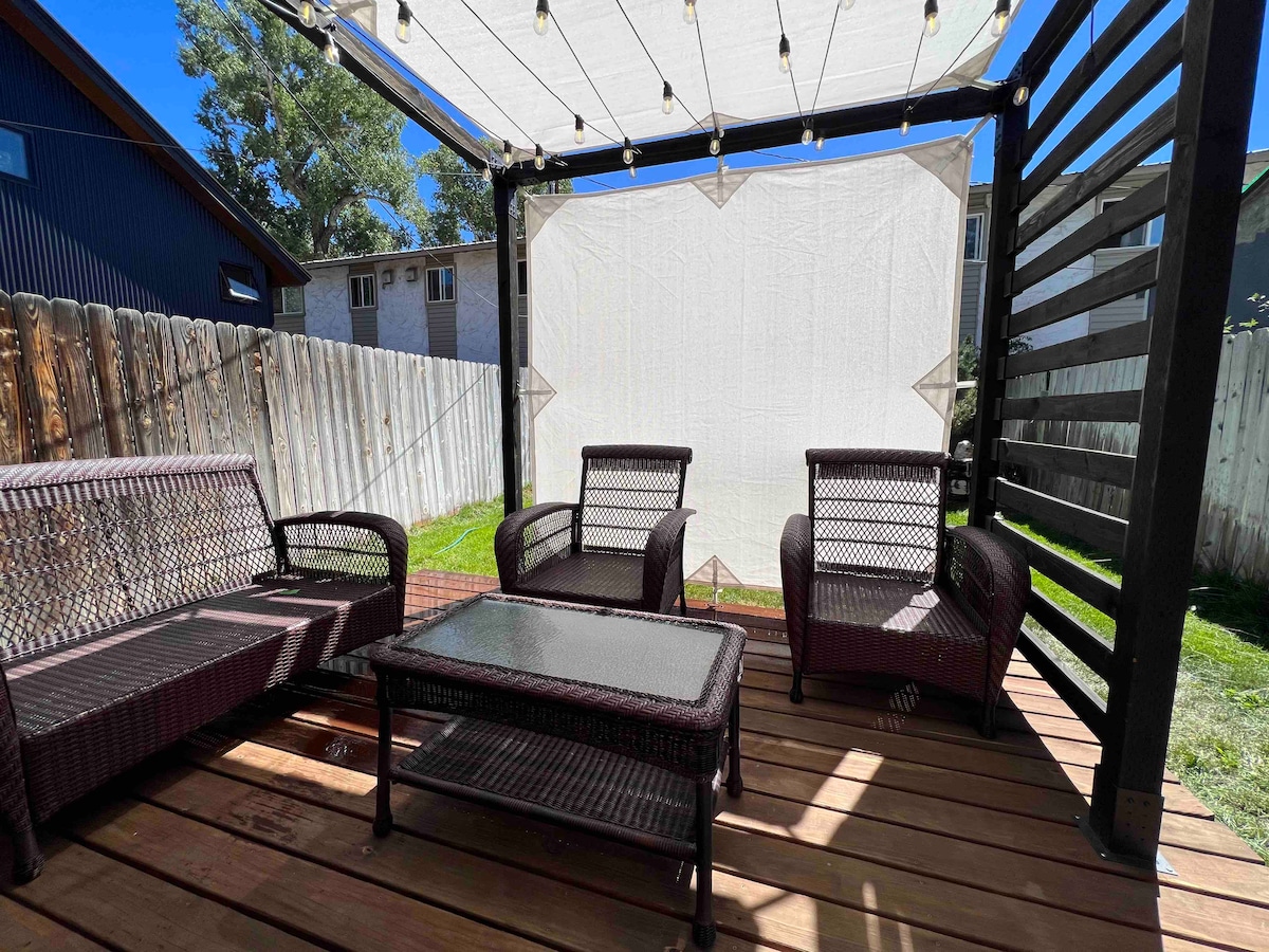 A shaded outdoor seating area is presented, featuring multiple wicker chairs and a table. String lights hang overhead, creating a welcoming ambiance. The space is enclosed by wooden privacy panels, with green grass visible in the background, providing a peaceful retreat.