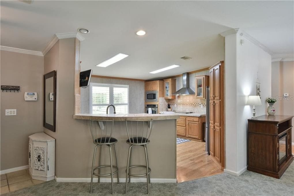 A light-filled kitchen area features an open layout, with a breakfast bar and two high-backed stools. Wooden cabinetry lines the walls, and modern appliances are visible. Soft, neutral colors are complemented by texture from the flooring and countertops, creating a welcoming ambiance.