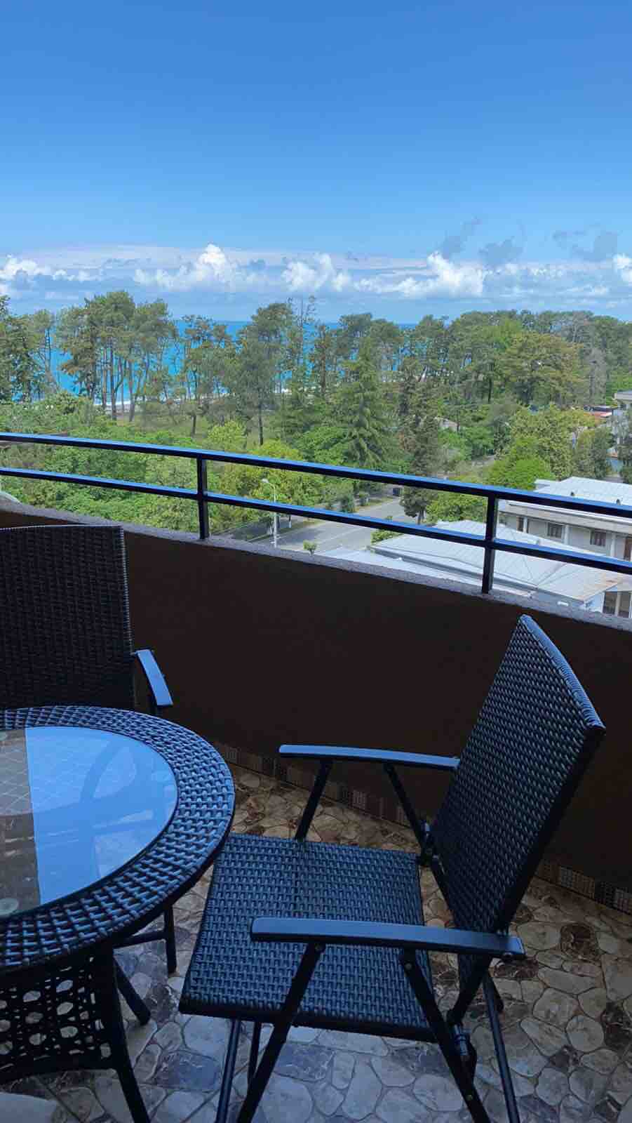 A balcony is shown, featuring a round glass table and two black wicker chairs. A view of the lush green trees extends towards a blue ocean, under a clear sky with fluffy clouds. The tranquil setting invites relaxation and enjoyment of the surroundings.