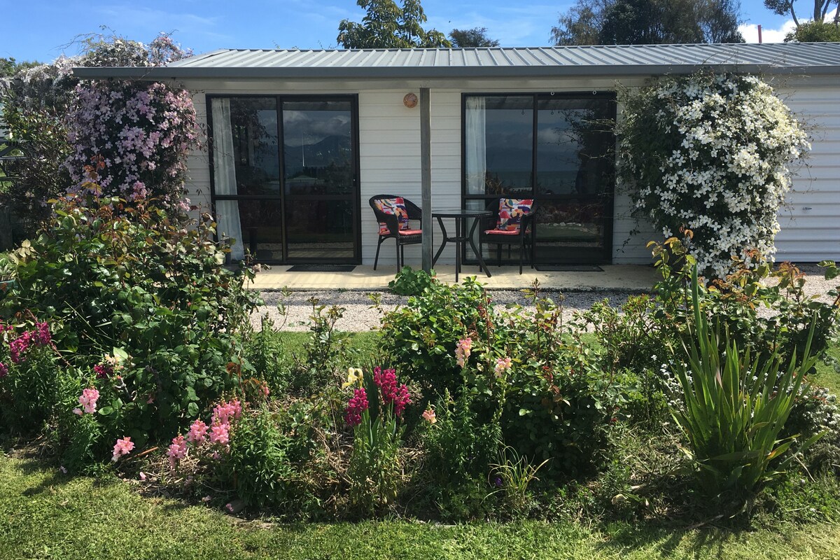 A charming exterior view of the accommodation is presented, featuring a well-maintained garden with colorful flowers. Two black chairs and a small table sit on the patio, framed by large glass doors that provide access to the interior.