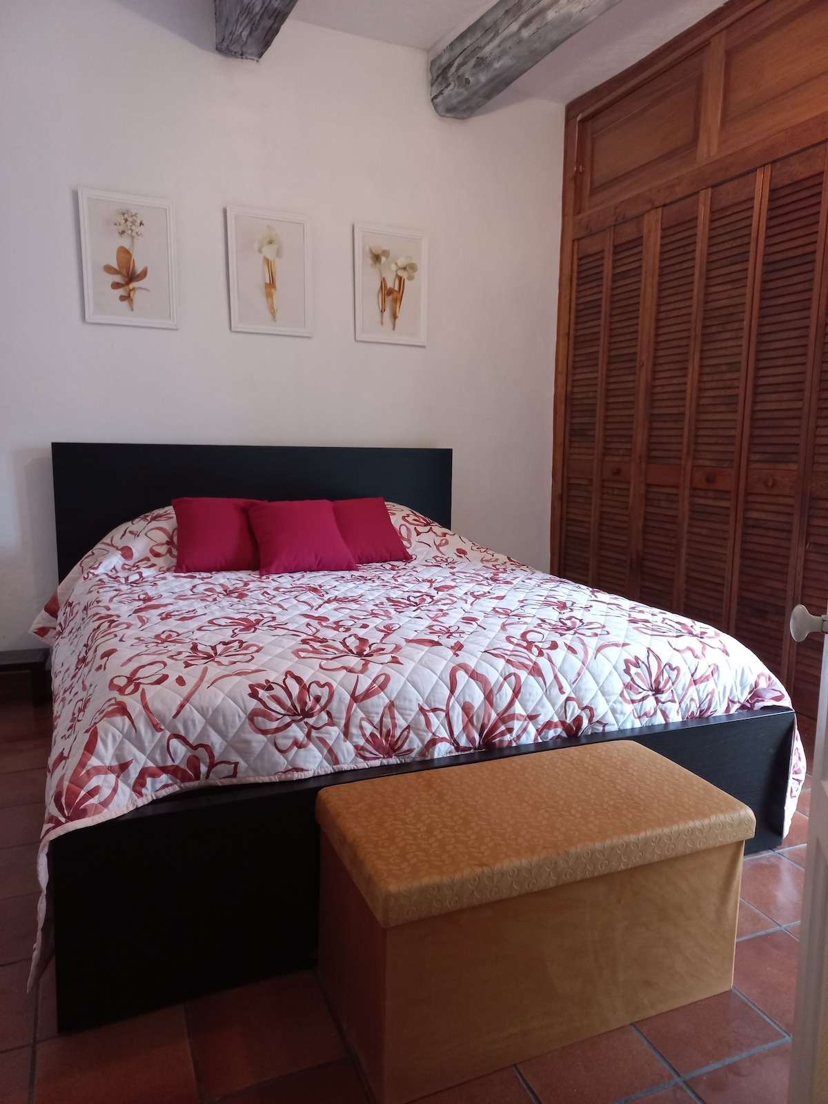 A spacious bed with a floral-patterned quilt is featured, complemented by two red accent pillows. Above the bed, three floral artwork pieces hang on the white wall. A wooden storage bench sits at the foot of the bed, adding functionality to the room.