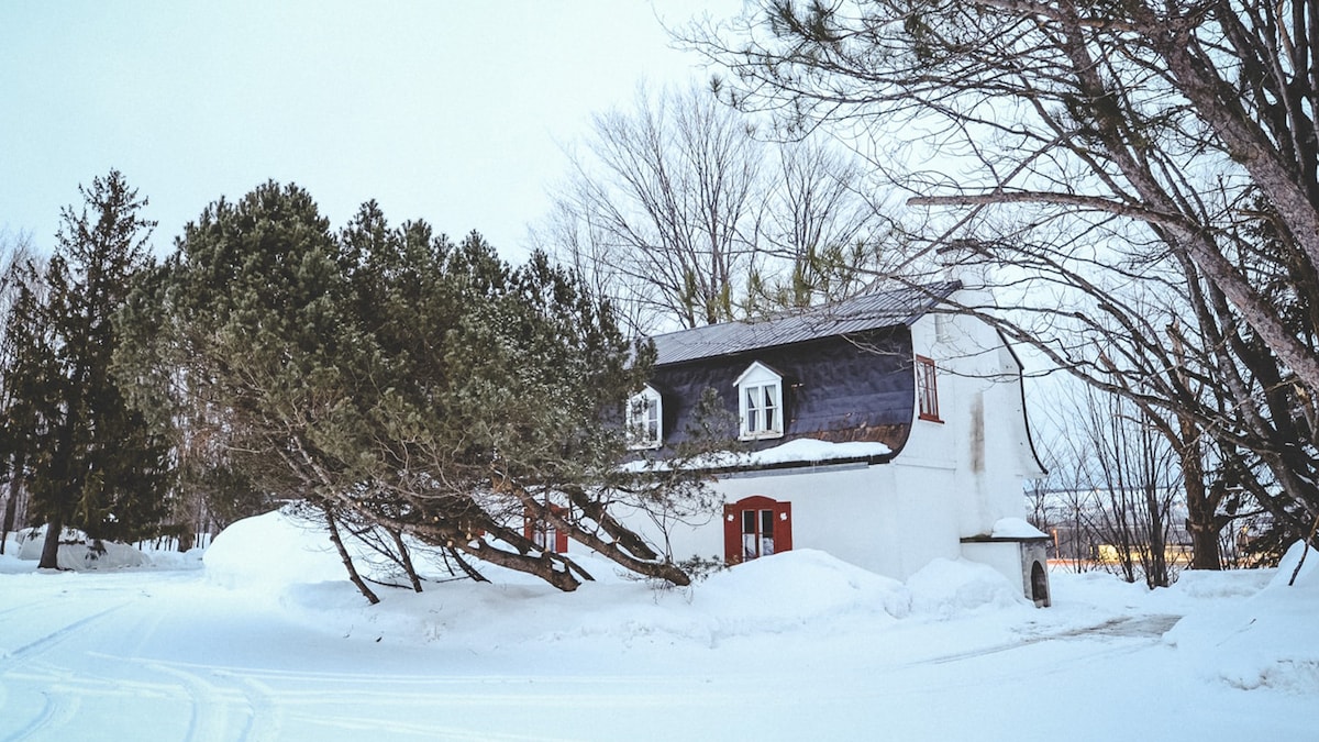 A quaint ancestral home is partially enveloped in snow, showcasing a white exterior contrasted with dark roofing and red window frames. Surrounding trees lean gently, and a serene winter landscape extends into the distance.