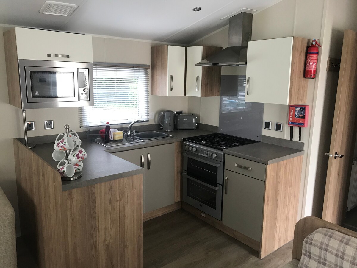 A modern kitchen area is equipped with essential appliances including a microwave, kettle, and stove. Light wood cabinetry contrasts with the sleek gray countertops. A set of white mugs is arranged neatly on the counter, while a window allows natural light to brighten the space.