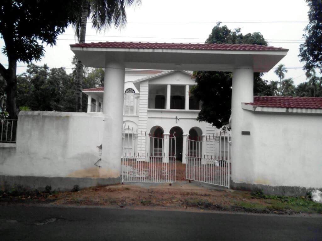 A grand entrance is framed by tall pillars, leading to a spacious farmhouse. A white gated fence provides access to the property, while lush greenery surrounds the building. The well-maintained exterior highlights the classic architecture of the heritage structure.