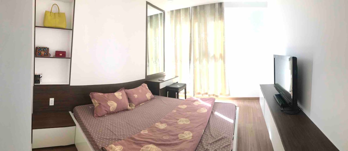 A well-lit bedroom features a spacious bed adorned with patterned bedding. A large mirror reflects natural light from the adjacent window, which is draped with soft, sheer curtains. A small bedside table is positioned next to the bed, and a television is mounted nearby.