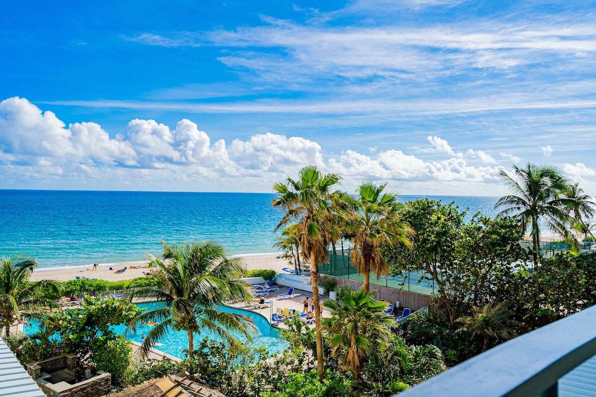A panoramic view showcases the sparkling ocean, sandy beach, and a lush pool area surrounded by palm trees. The sky features a mix of white clouds and bright blue tones, enhancing the beachfront atmosphere. Lounge chairs are visible near the pool, inviting relaxation.