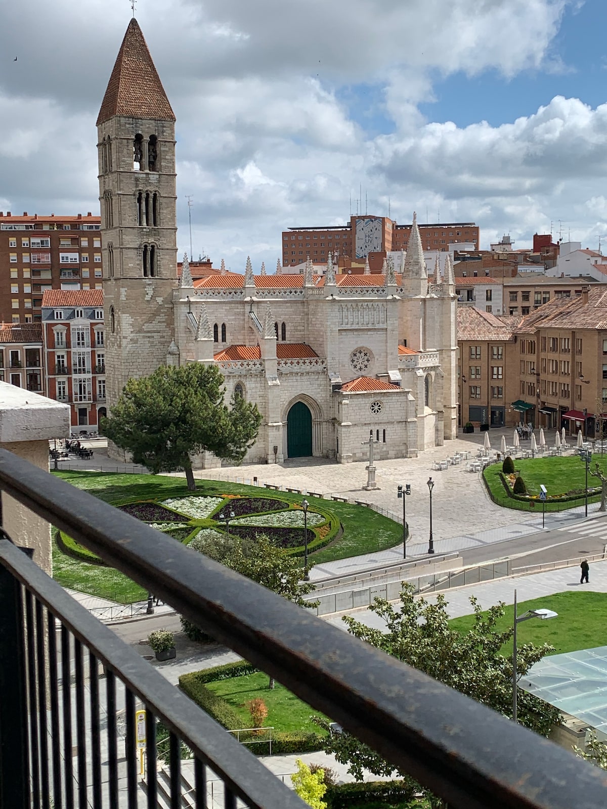 The image captures a view of the historic Church of Santa María de La Antigua, featuring a tall bell tower and intricate stonework. Lush green spaces and neatly arranged gardens are visible in the foreground, framed by a balcony railing that enhances the scenic perspective.