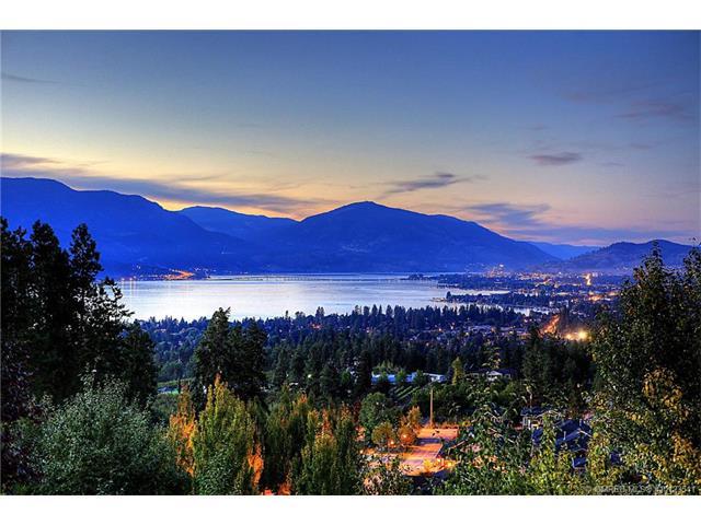 A panoramic view captures the serene landscape at dusk, showcasing shimmering lake waters bordered by mountains. The softly illuminated city can be seen in the distance, framed by trees and lush greenery in the foreground.