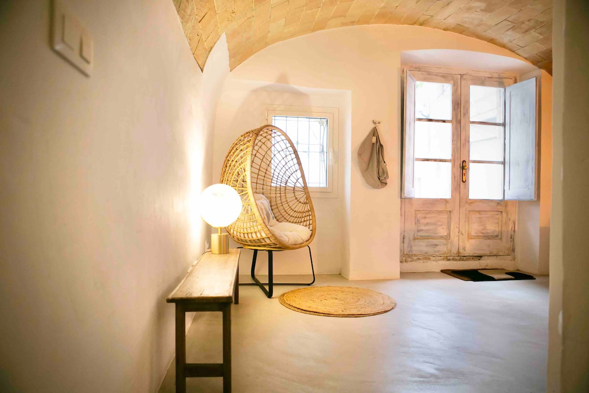 A welcoming entryway is presented with a unique woven chair positioned next to a rustic door. Natural light filters through window panes, illuminating the soft, neutral tones of the space. A round rug lies beneath the chair, enhancing the inviting atmosphere.