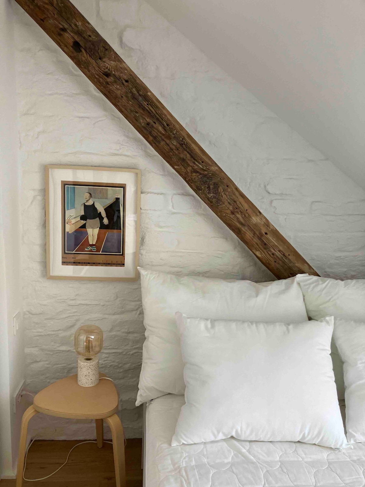 A cozy sleeping area is presented with a white bed adorned with large, soft pillows. A minimalist bedside table holds a unique lamp, while a framed art piece featuring a person stands against a textured white brick wall, complemented by exposed wooden beams.