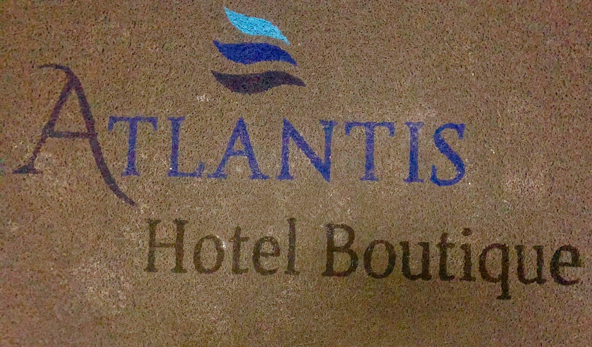 The image displays the logo of Atlantis Hotel Boutique, featuring elegant typography in blue and black against a textured background. The design conveys a contemporary aesthetic, with wave motifs suggesting a connection to water and relaxation.