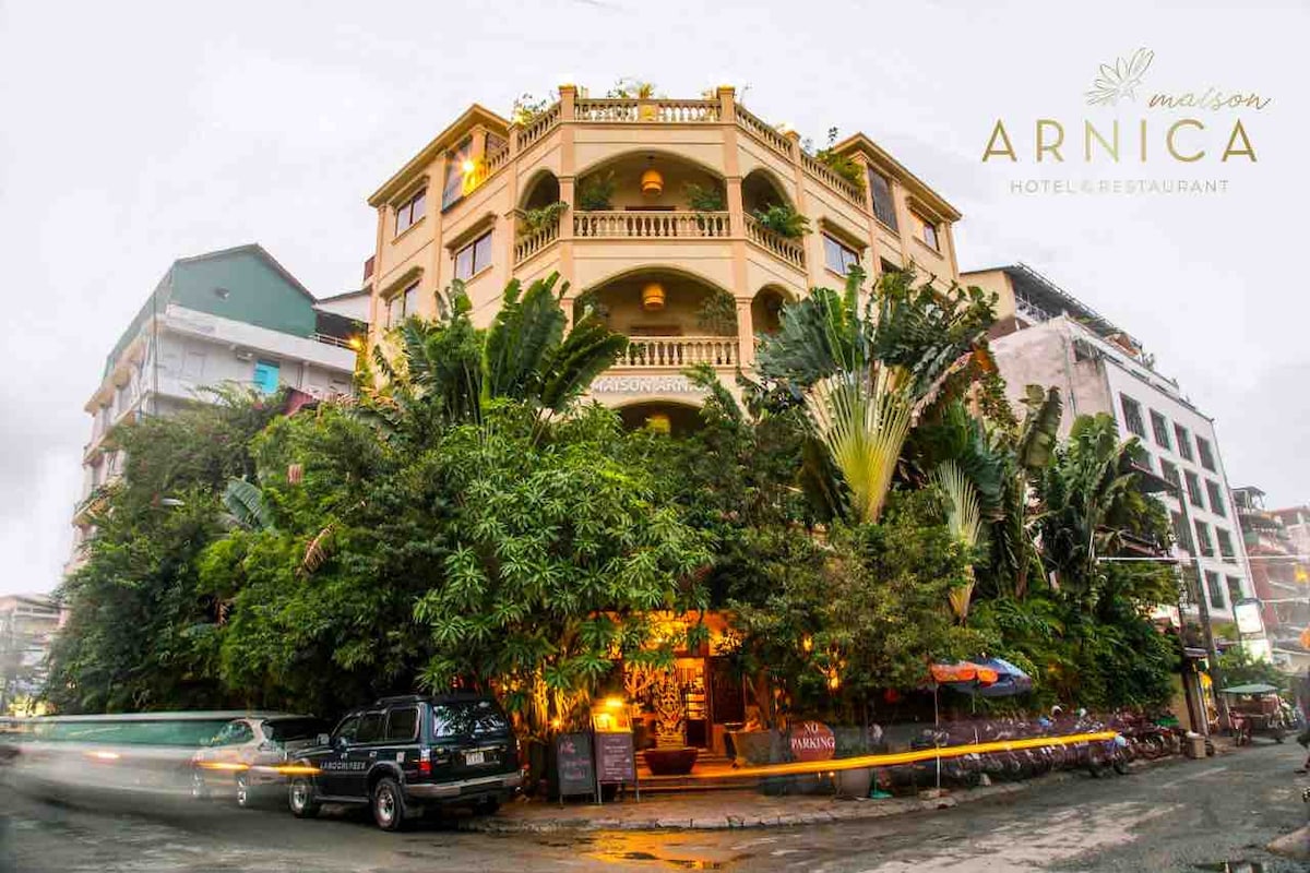 A charming hotel building is surrounded by lush greenery, displaying multiple balconies and a welcoming entrance. Soft lighting from the ground-level area highlights the landscaping and creates a warm ambiance. The structure is centrally located, with visible nearby buildings partially obscured by foliage.