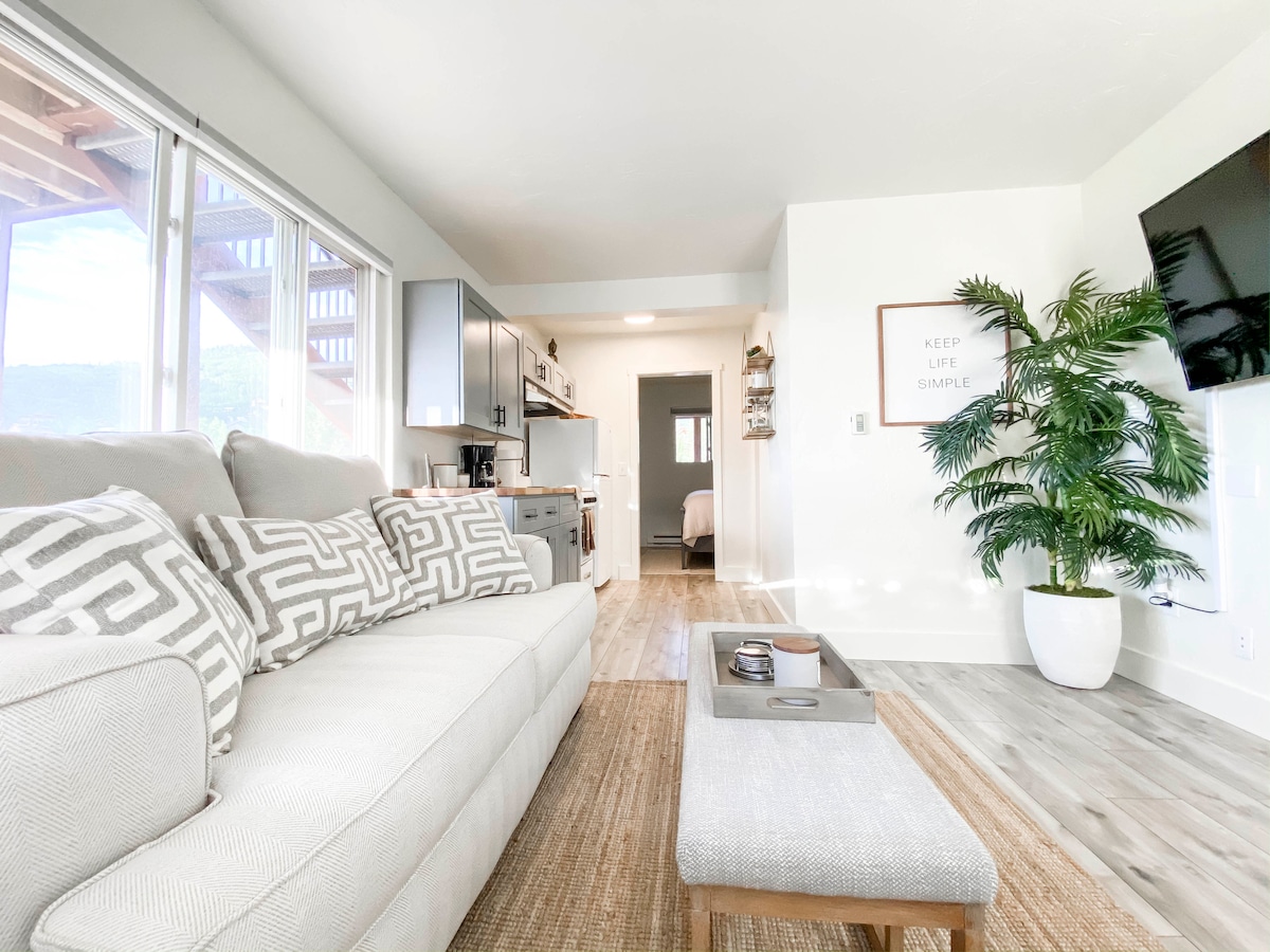 A spacious living area features a light-colored sectional sofa adorned with geometric throw pillows. A minimalist coffee table sits in front, and large windows provide natural light and views of the outdoor space. A potted plant adds a touch of greenery, enhancing the inviting ambiance.