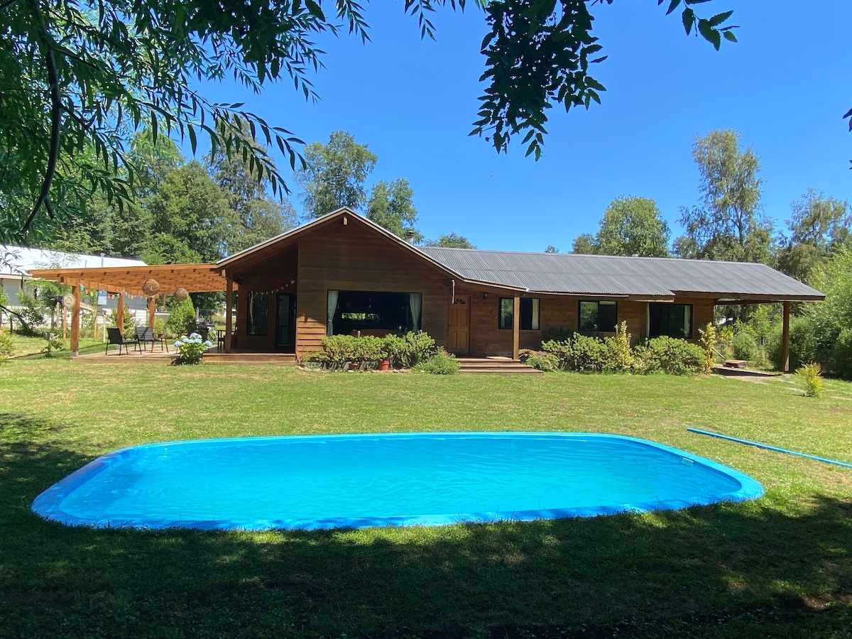 A wooden house is set against a clear blue sky, surrounded by lush greenery. A tranquil blue pool is positioned in the foreground, offering a refreshing spot for relaxation. Large trees provide shade, enhancing the serene outdoor environment.