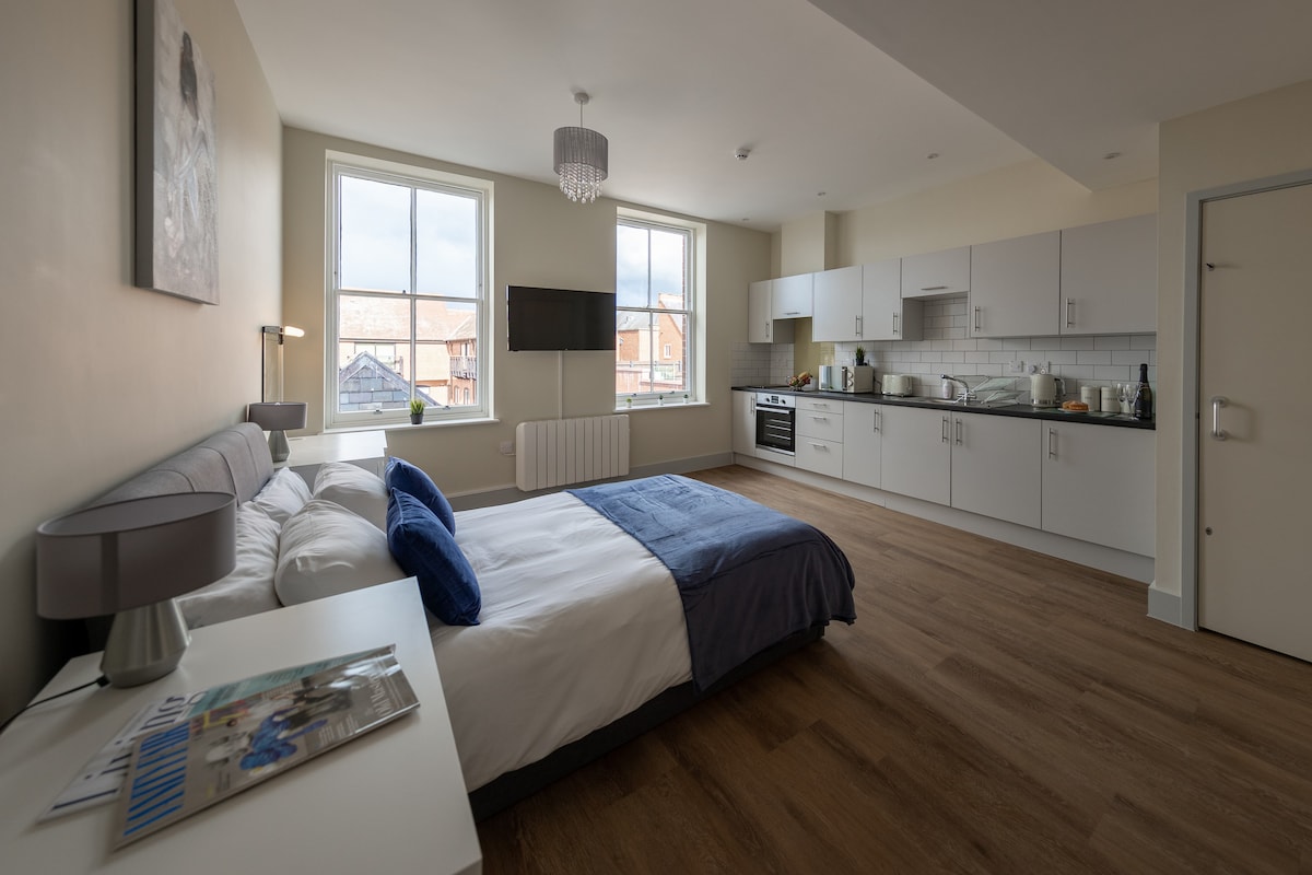 An open-plan studio features a spacious sleeping area with a double bed dressed in neutral linens and accented by blue pillows. A flat-screen TV is mounted on the wall. The modern kitchen includes sleek cabinetry and integrated appliances, with windows allowing natural light to fill the room.