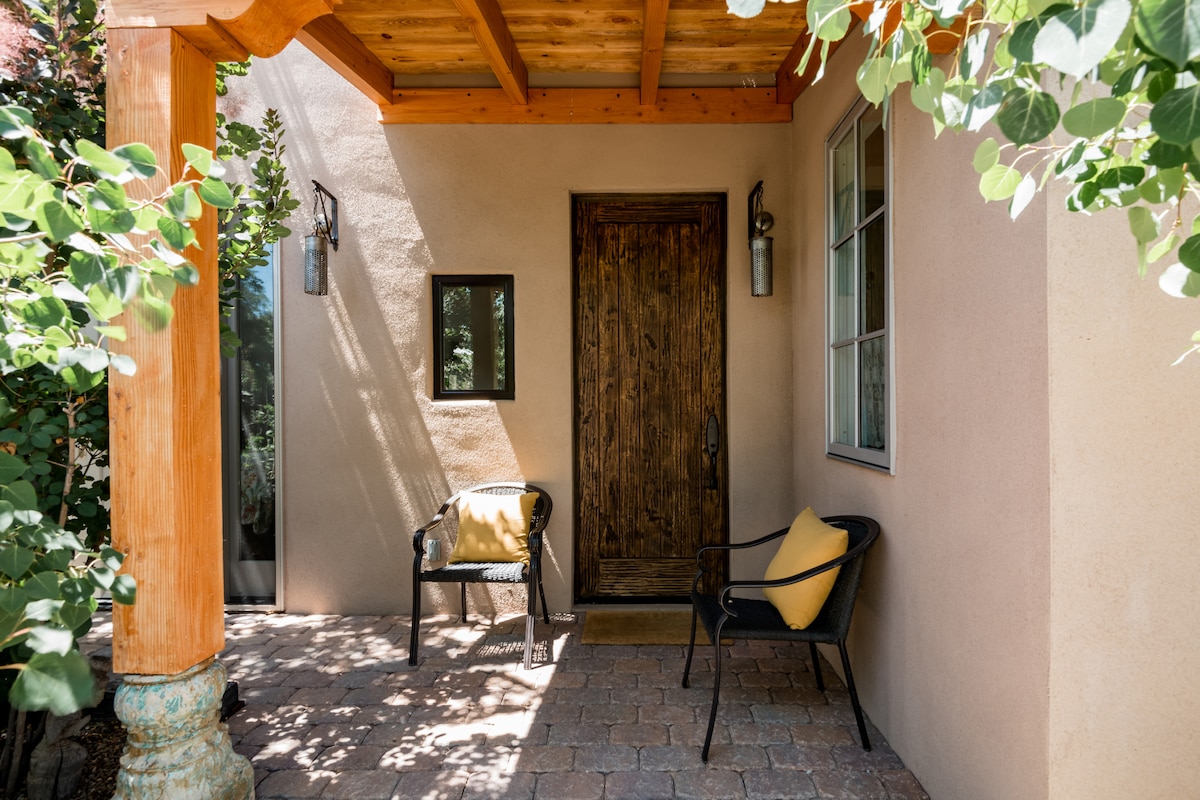 A welcoming entryway is characterized by a rustic wooden door framed by a light-colored stucco wall. Two black chairs with yellow cushions are positioned under a wooden overhang, surrounded by lush greenery. Natural light filters through nearby windows, enhancing the inviting atmosphere.