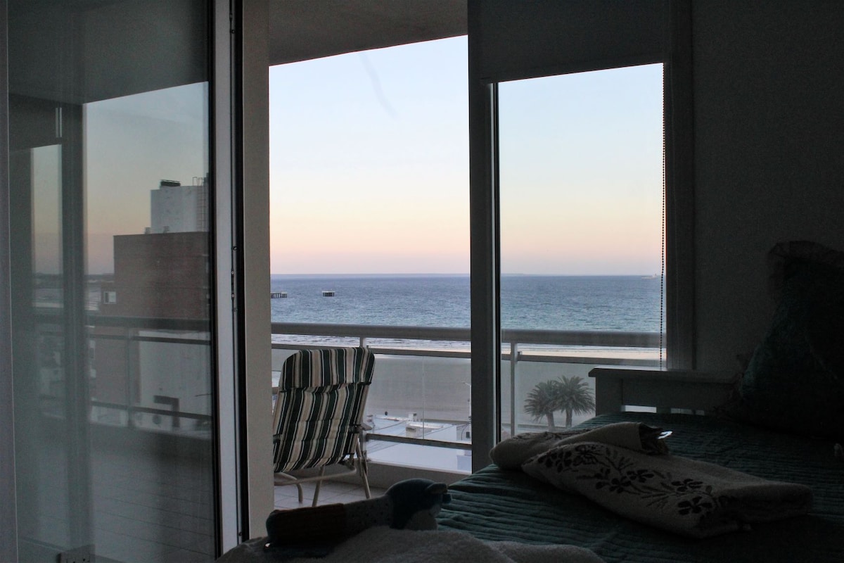 A view from the bedroom showcases a serene ocean horizon, framed by large glass doors. The early evening sky transitions in color, while a pair of outdoor chairs is visible on the balcony. Light filters softly into the interior space.