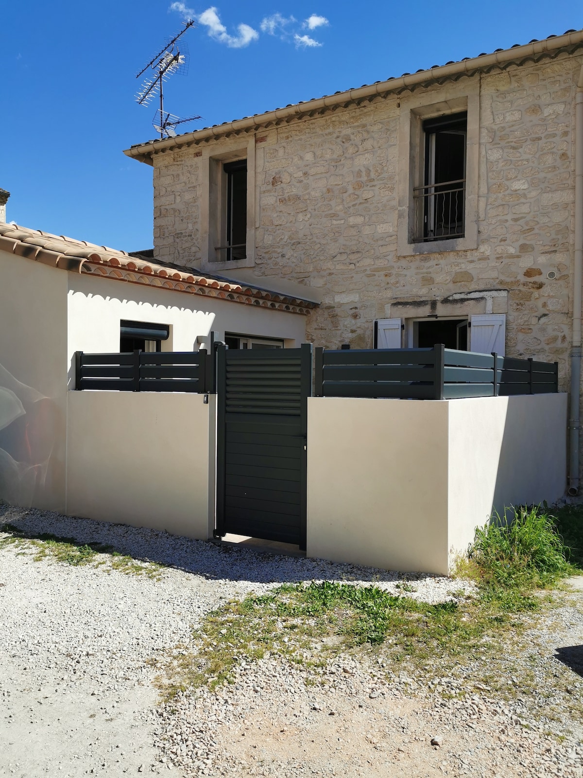 A private outdoor area is enclosed by a modern privacy fence, featuring a secure gate. The background showcases a stone house with wooden shutters, while gravel and small patches of grass surround the space, contributing to a serene and secluded environment.
