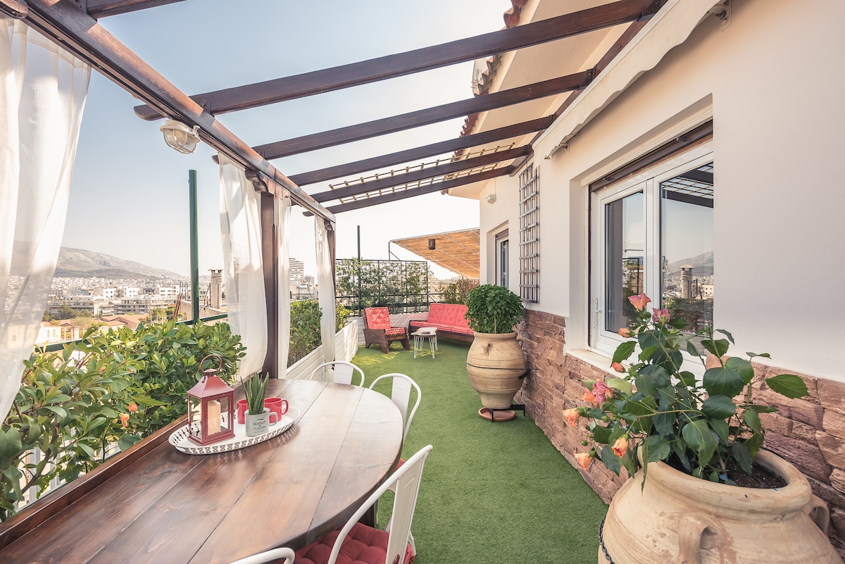 A private terrace offers a panoramic view, featuring a spacious dining table surrounded by greenery. Sunlight bathes the area, highlighting comfortable seating arrangements adorned with cushions. Elegant terracotta planters enhance the space with vibrant flowers, while curtains billow gently with the breeze.