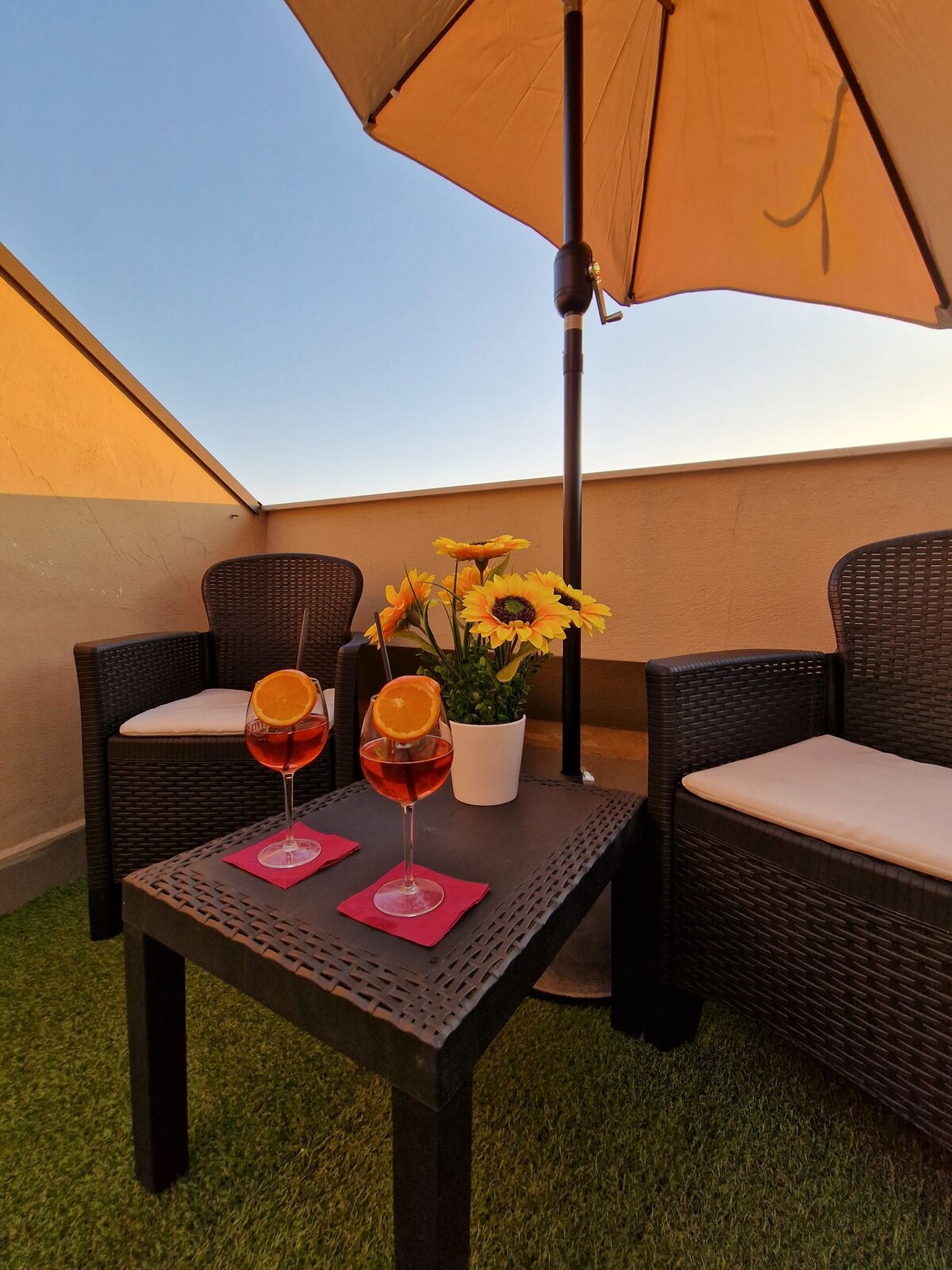 A relaxing outdoor seating area features two wicker chairs and a small table topped with two drinks garnished with orange slices. A potted plant with yellow flowers adds a touch of color. A large umbrella provides shade, set against a clear blue sky.
