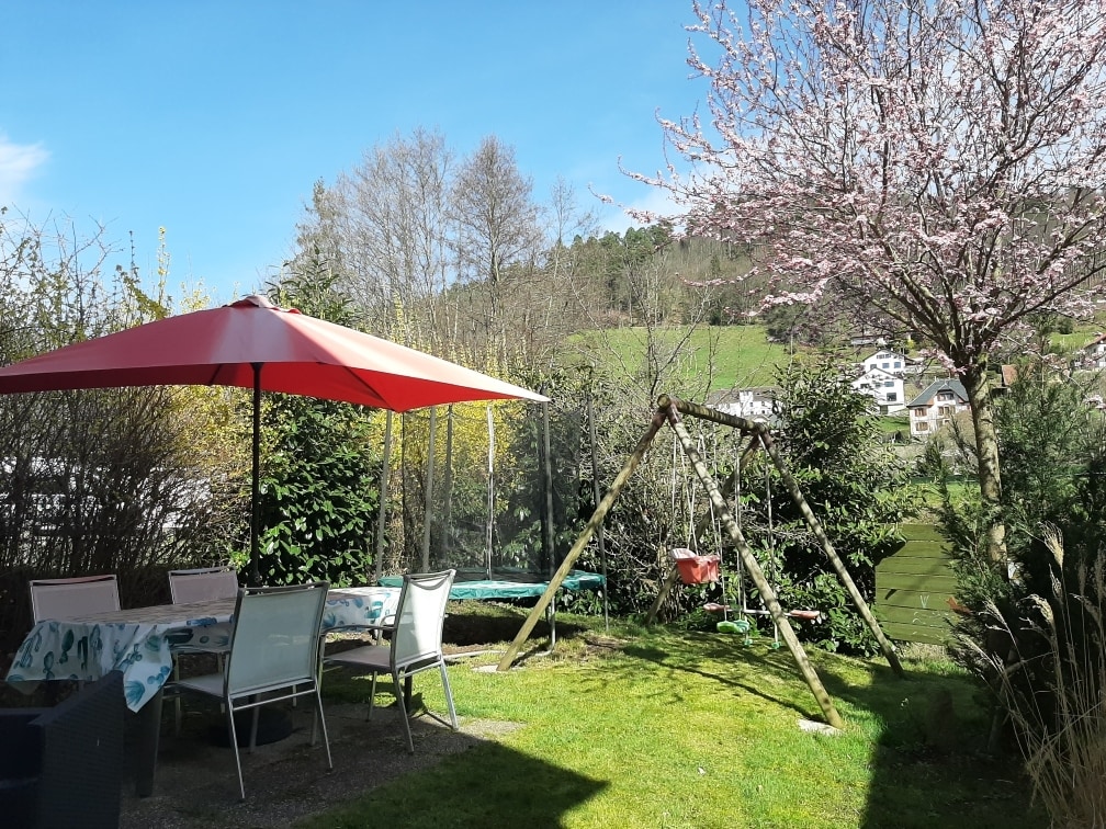 A private garden features a shaded patio area with a red umbrella over a table and chairs. A swing set is visible nearby, along with a trampoline. Lush greenery and blooming trees provide a backdrop, complemented by a scenic hill in the distance.