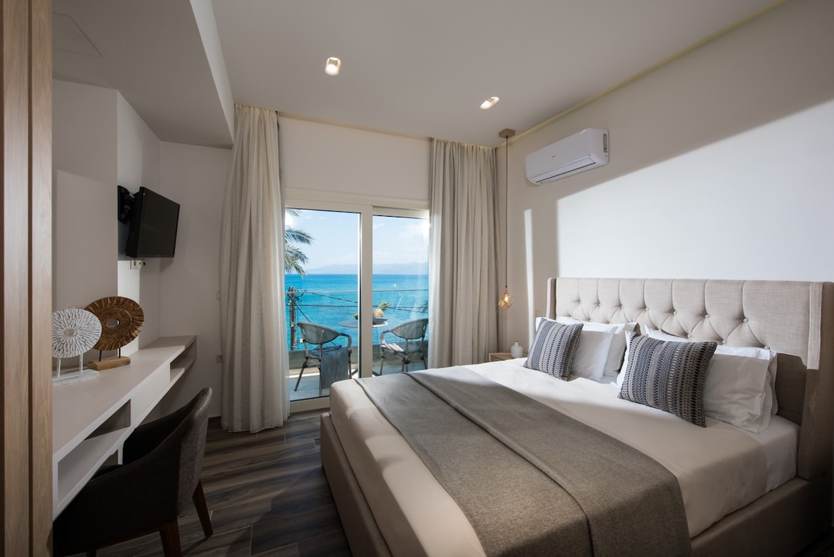 A contemporary bedroom features a spacious bed adorned with decorative pillows and a soft throw. A desk and chair are positioned nearby. Large glass doors open to a balcony with sea views, framed by light-colored curtains allowing natural light to fill the room.