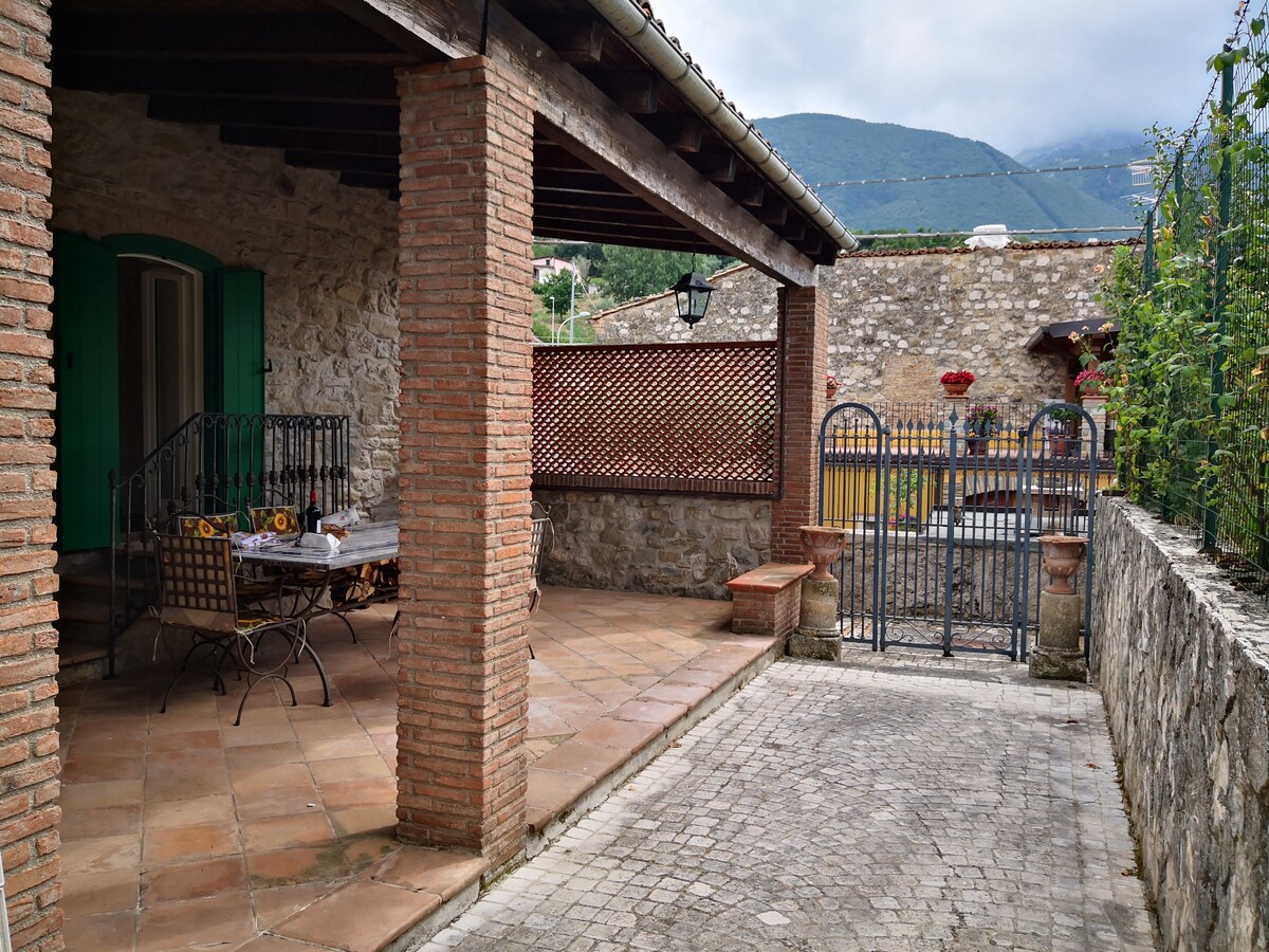 An outdoor patio area features a stone wall, terracotta flooring, and a wooden beam ceiling. A table is set for dining, complemented by surrounding greenery. A wrought iron gate leads to a picturesque view beyond, with mountains visible in the background.