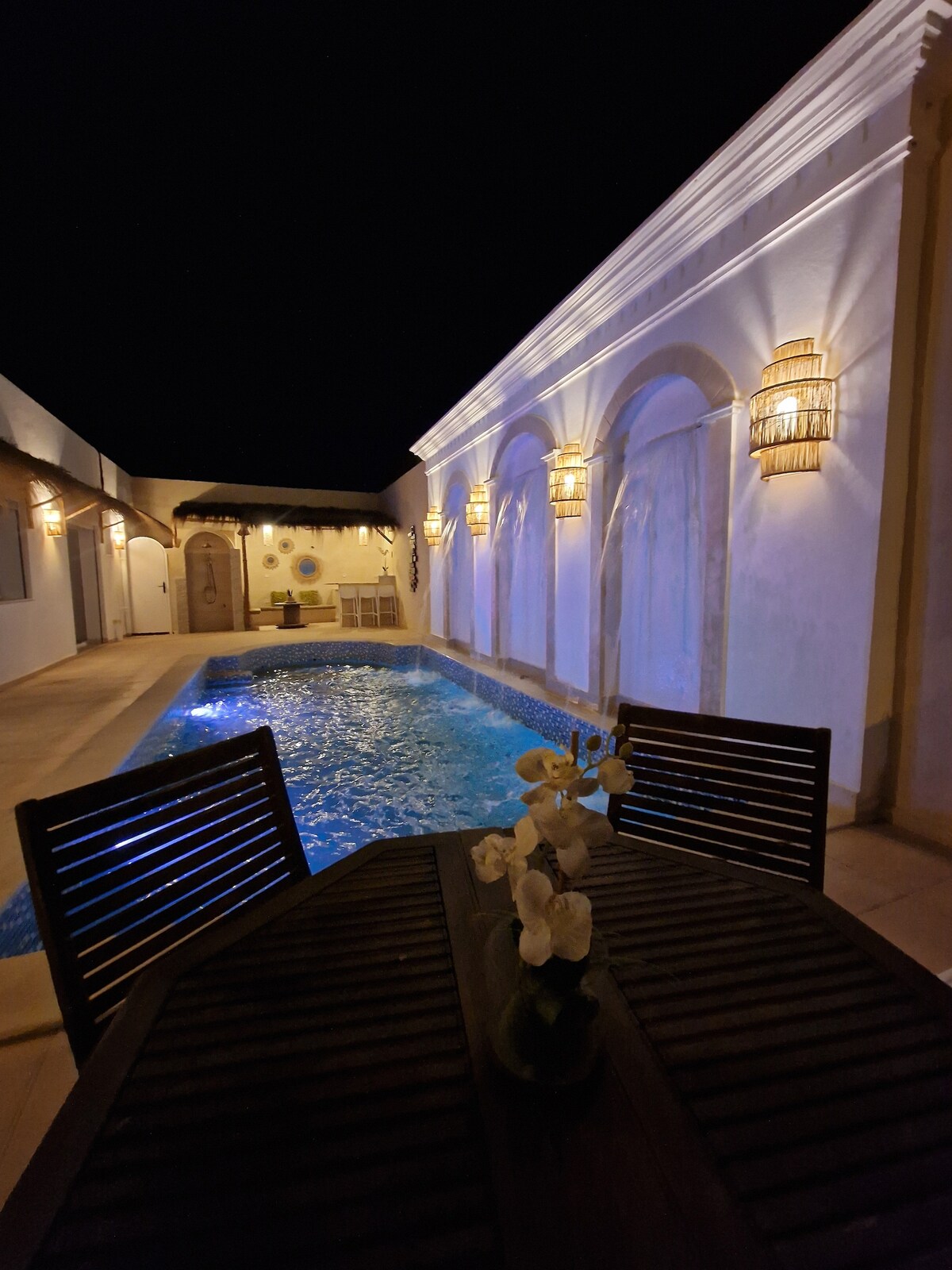 A tranquil outdoor space features a swimming pool illuminated in blue against a backdrop of softly lit walls. A wooden table with a vase of flowers is positioned nearby, inviting relaxation. Surrounding arches and lanterns add to the serene atmosphere in the evening setting.