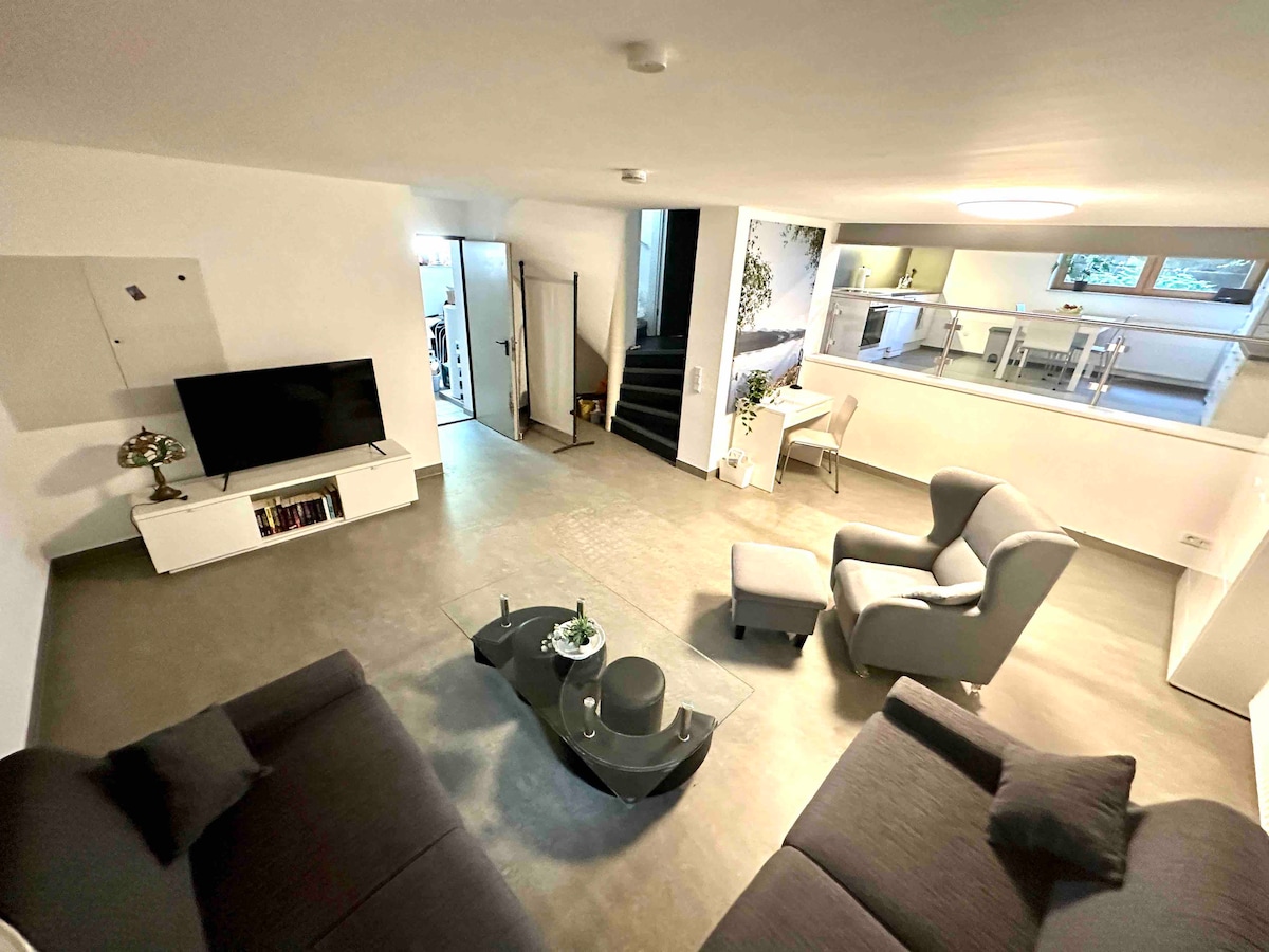 The spacious living area features a large flat-screen TV and modern furnishings, including a gray armchair and a dark sectional sofa. Natural light filters in through windows, complementing the minimalistic decor and open layout that leads into the kitchen area.