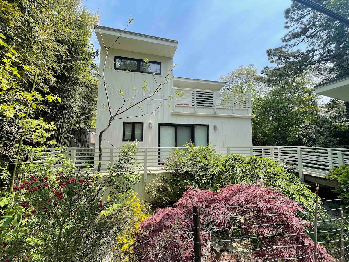 A modern-style house is surrounded by vibrant greenery and colorful plants. Its multi-level design features expansive windows and a balcony on the second floor. A wooden walkway leads to the entrance, creating a connected feel with the natural landscape.