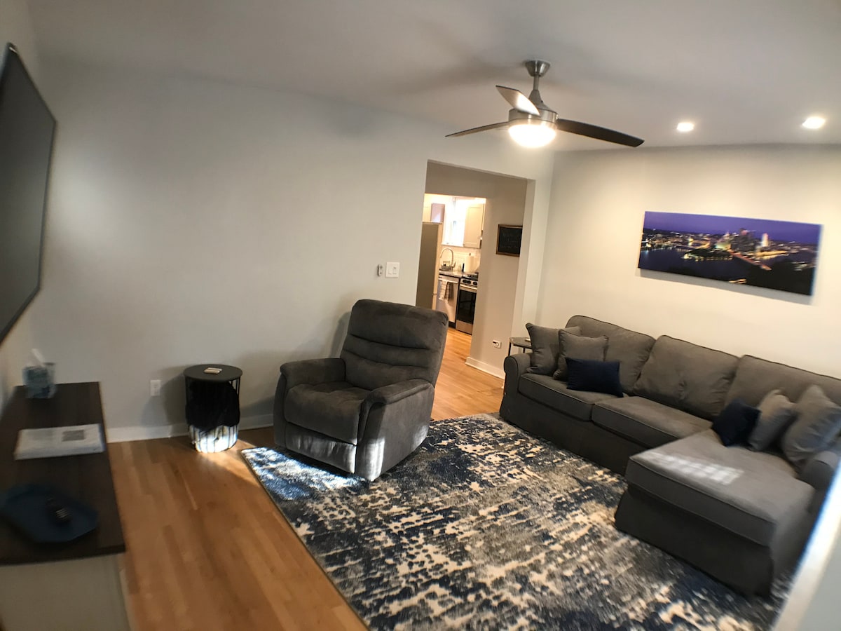 A living room is depicted with a gray sectional sofa and a reclining chair, arranged around a low coffee table. A large television is mounted on the wall opposite. A rug with blue tones adds warmth to the space, while soft lighting is provided by a ceiling fan.