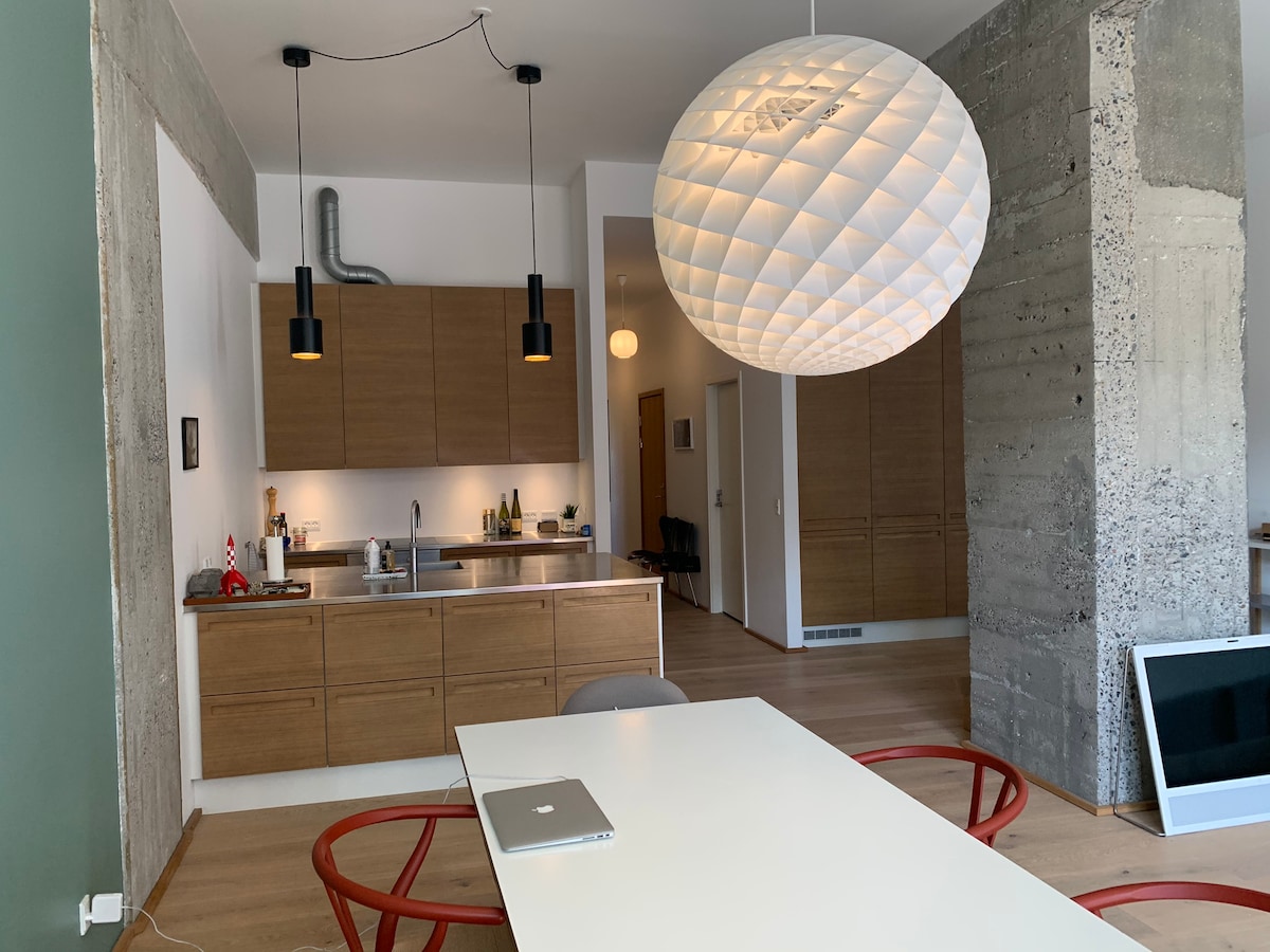 A modern dining area features a light-colored table surrounded by red chairs. A large pendant light hangs above, providing bright illumination. The kitchen area is visible in the background, showcasing wooden cabinets and sleek appliances. Concrete walls add a contemporary touch to the space.