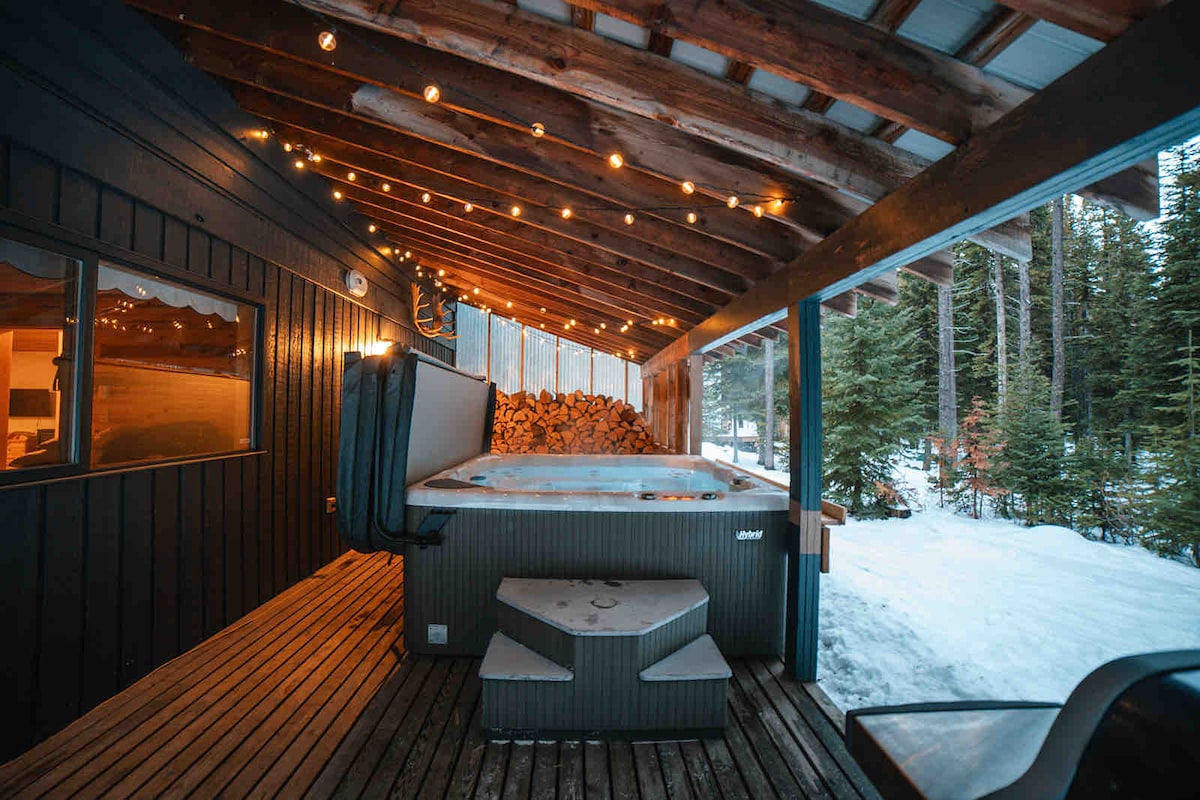 A covered outdoor area features a hot tub surrounded by wooden decking and fairy lights that add a warm touch. Behind the tub, a stack of firewood is visible, and a snow-covered landscape is observed beyond the structure.