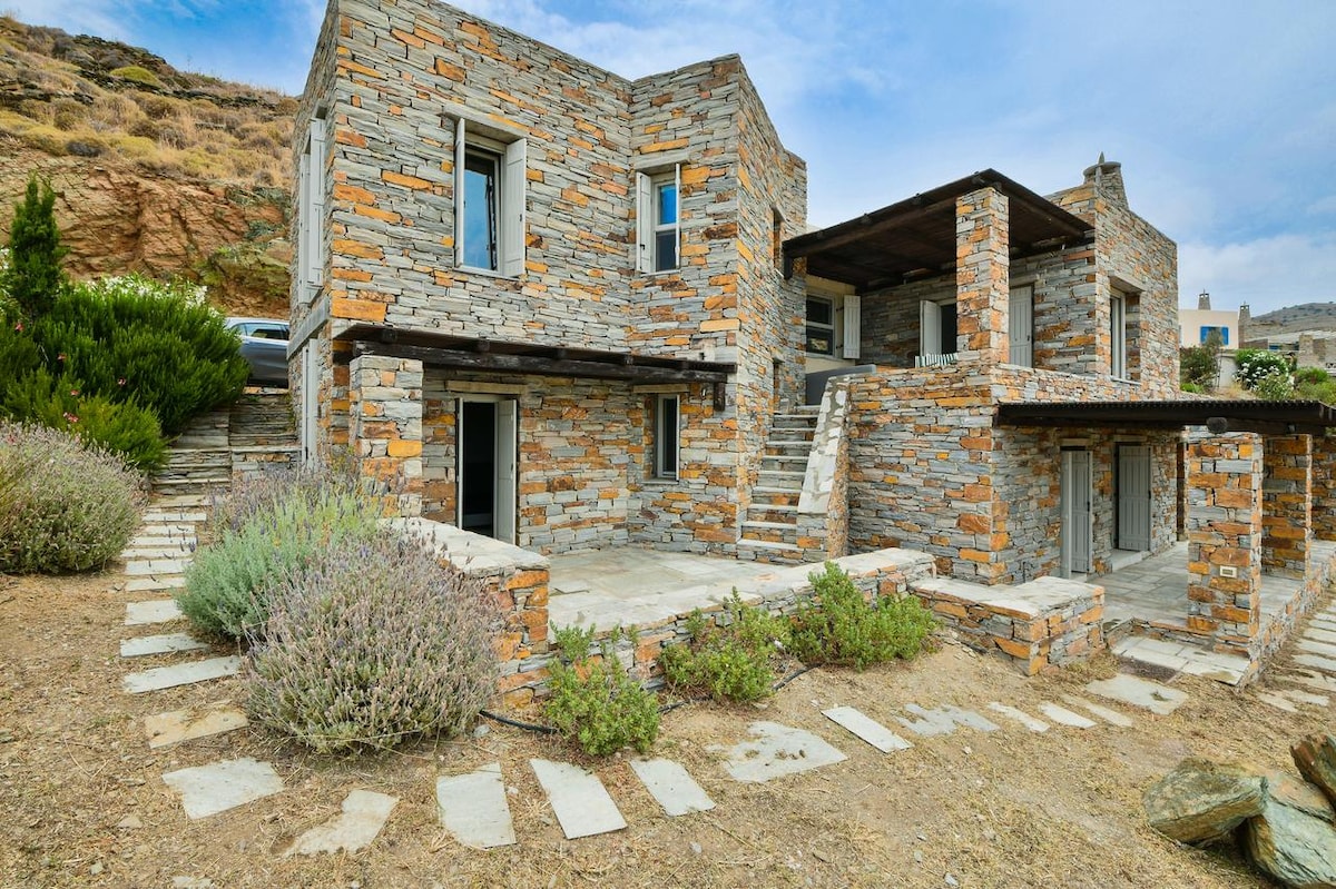 A rustic stone villa features a sturdy facade with a blend of gray and orange hues, complemented by large windows. Surrounding the structure, a well-maintained pathway leads through patches of greenery, enhancing the natural setting against a backdrop of rocky terrain.